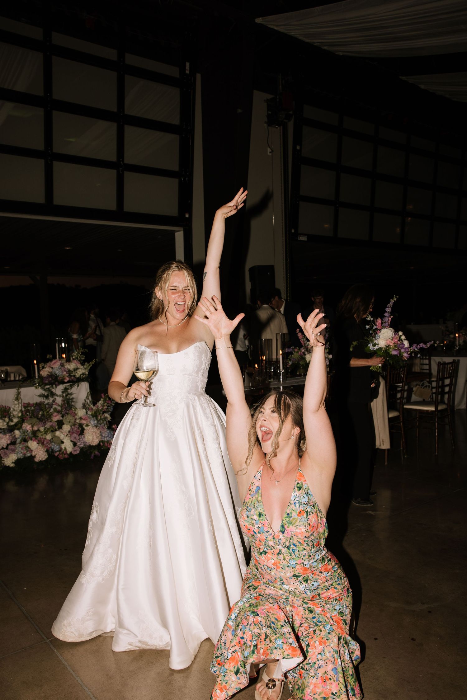 Excited bride in white strapless gown celebrates with guest in floral dress on wedding reception dance floor.