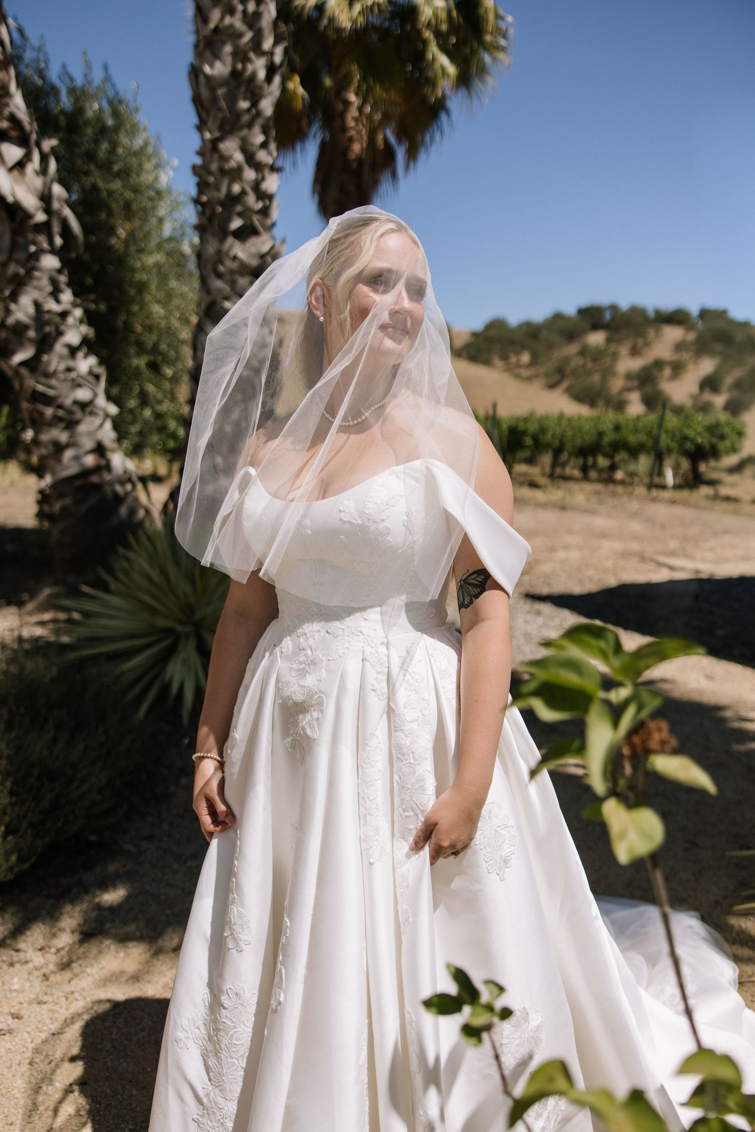 Blonde bride in white gown with veil at sunny vineyard wedding.