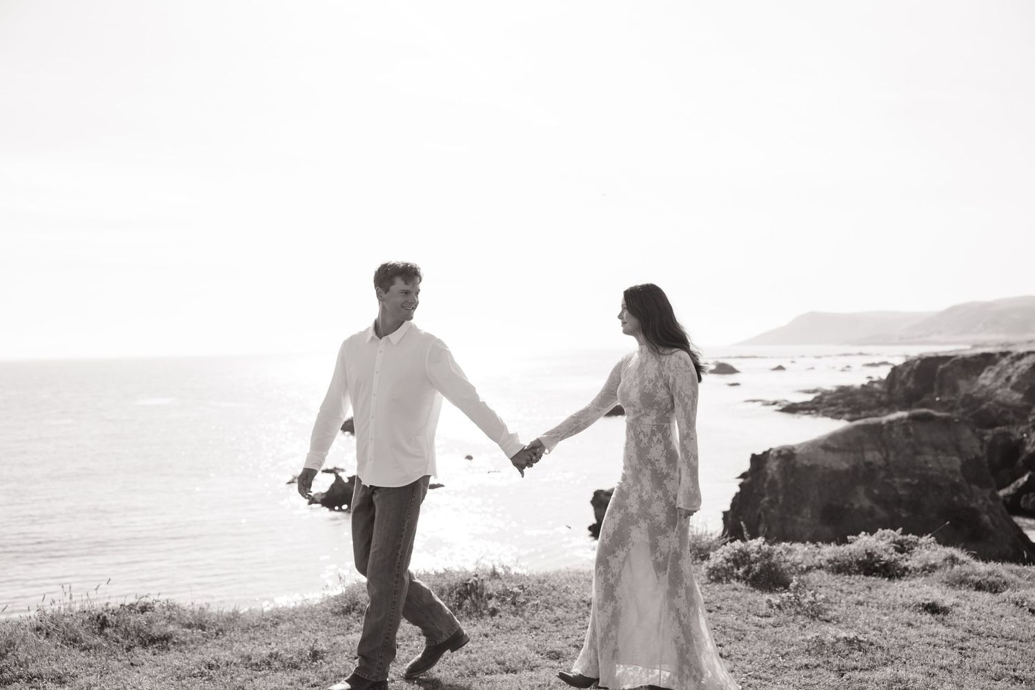 Couple walking hand in hand along coastal cliffside, black and white wedding photography.