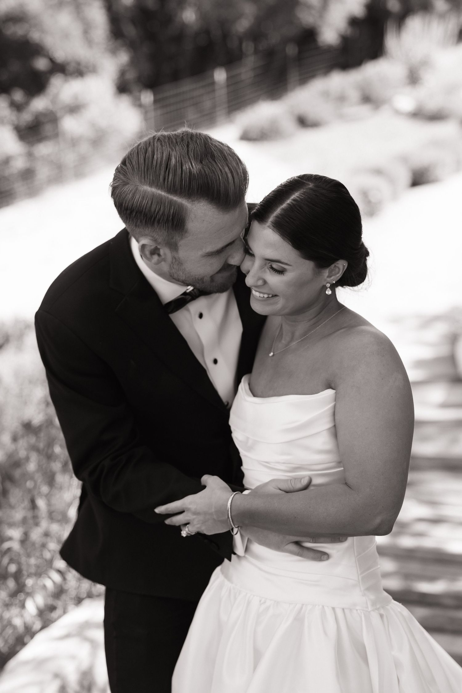 Black and white photo of groom in tuxedo tenderly embracing smiling bride in strapless wedding gown.
