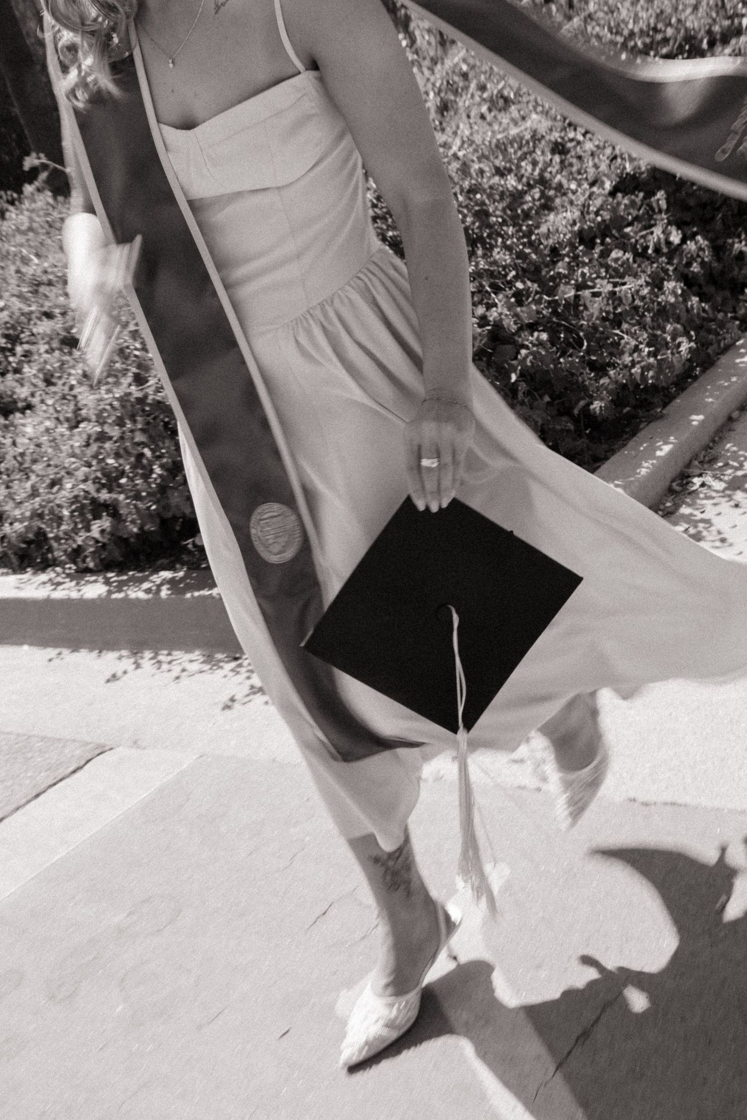 Black and white photo of a woman in a white dress holding a graduation cap while walking down outdoor steps.