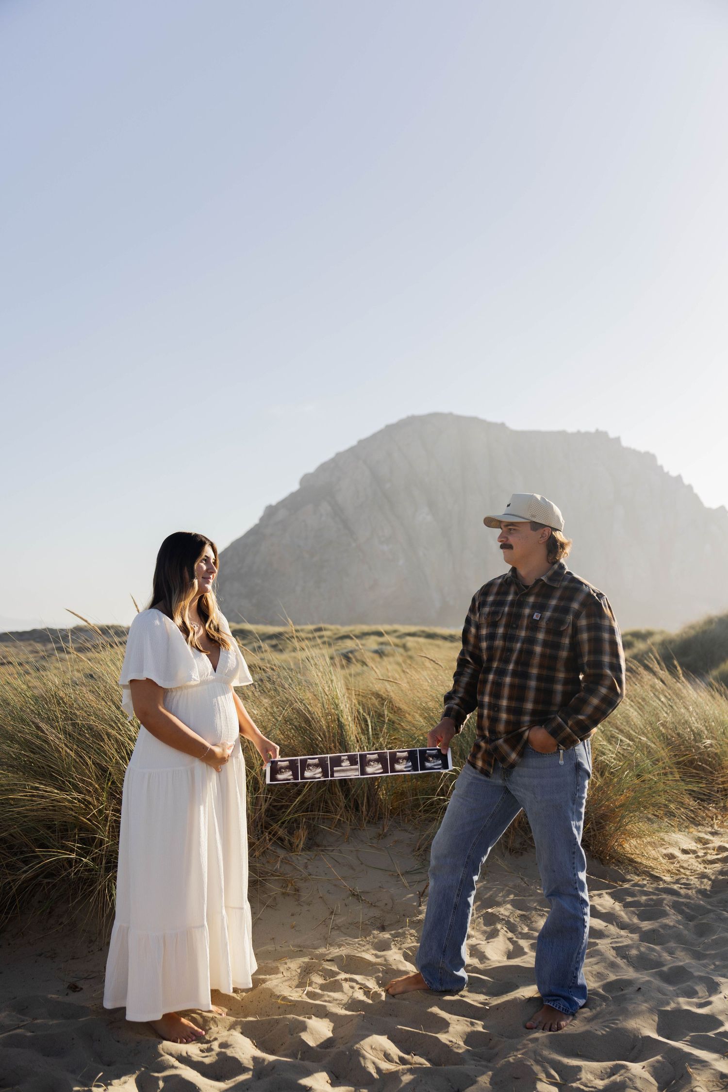 Pregnant woman in white dress with partner holding ultrasound strip on sandy beach near Morro Rock.