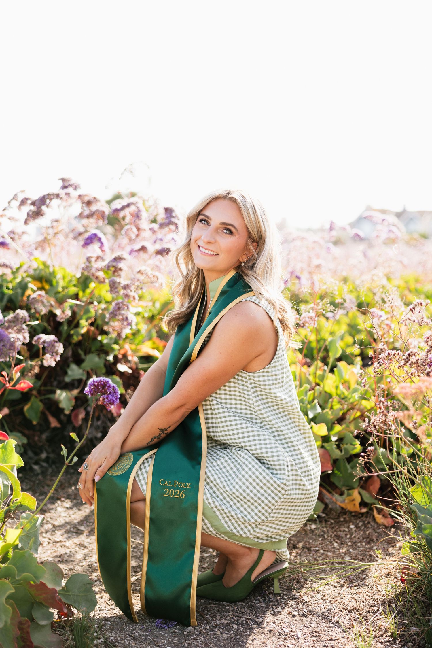 Smiling blonde woman in graduation stole kneeling in flower garden for Cal Poly 2026 senior photos.