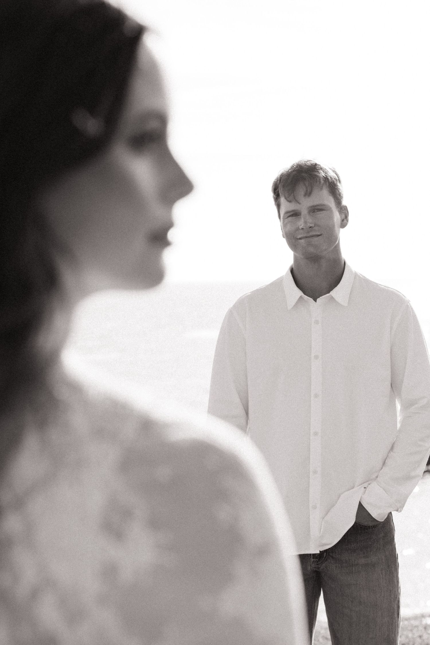 Black and white photo of man in white shirt gazing at woman in lace dress by ocean.