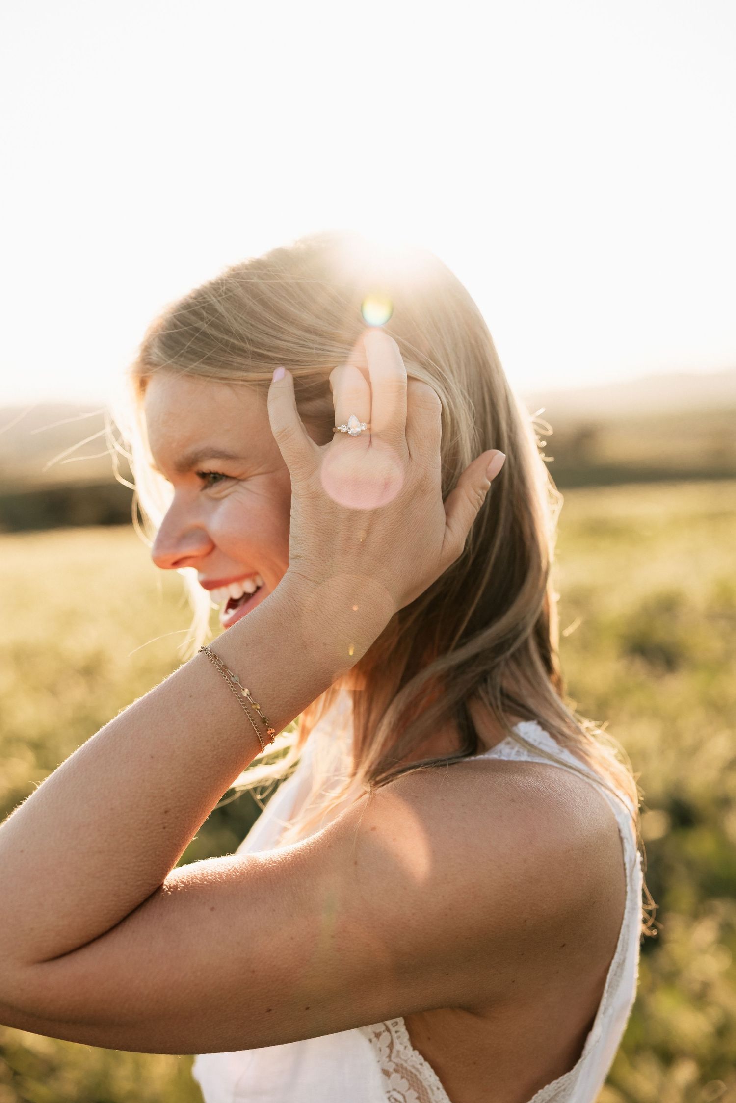 Smiling blonde woman in white dress showing engagement ring in golden sunlight.