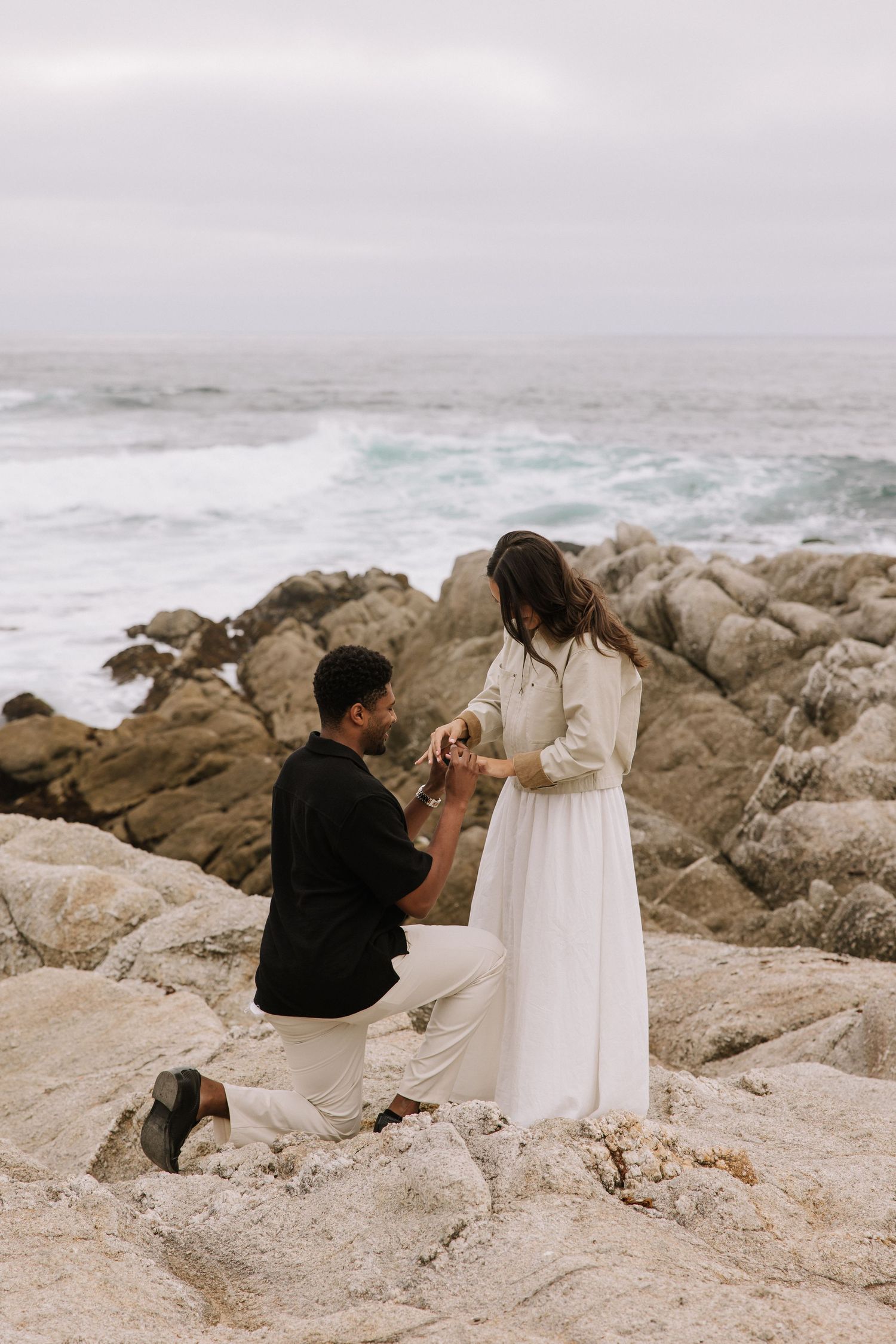 Man kneeling on rocky coastal shore proposing to woman in white dress with ocean waves behind them.