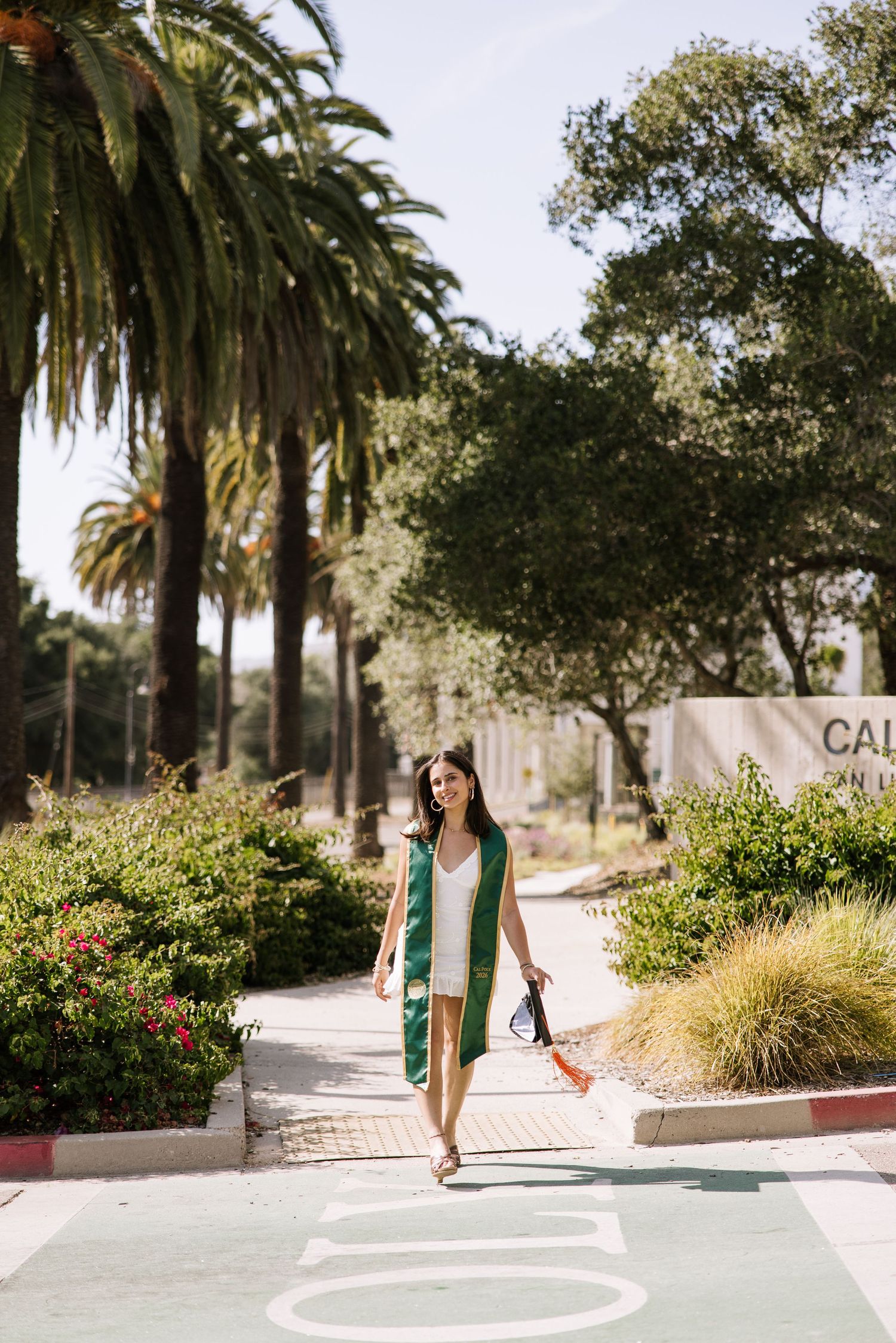Smiling graduate in white dress and green stole walking along sunny campus path with palm trees.