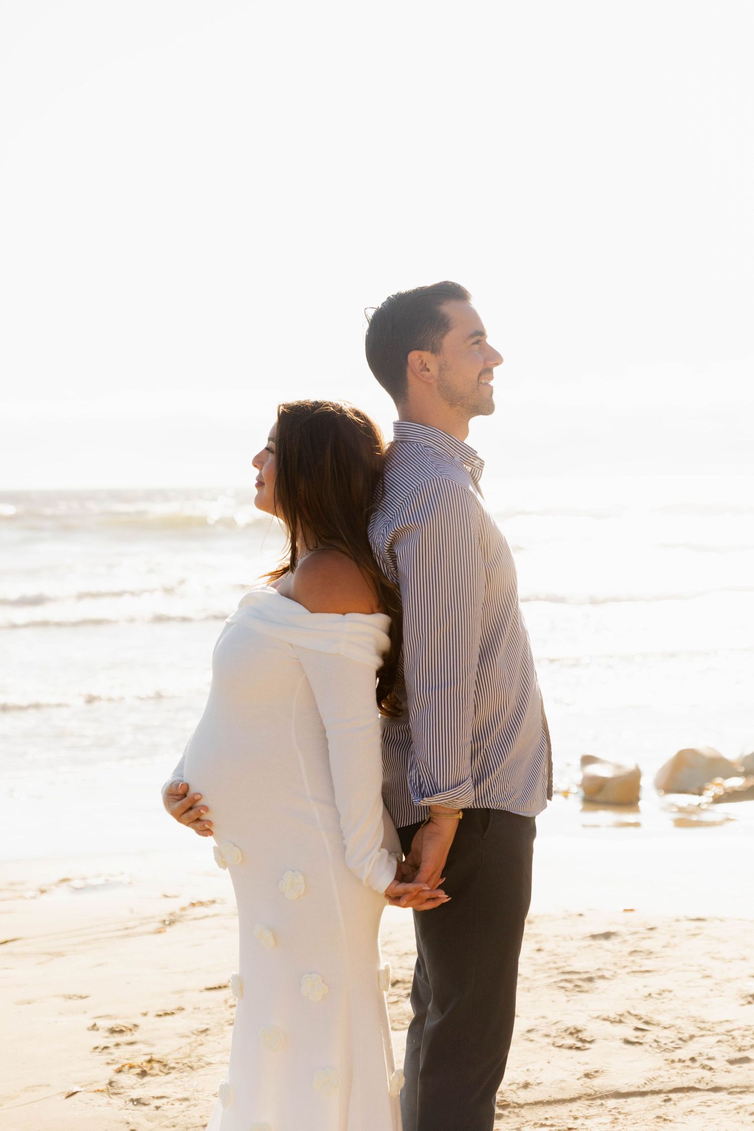 Smiling pregnant couple standing back to back on sunny beach, woman in white floral maternity dress.
