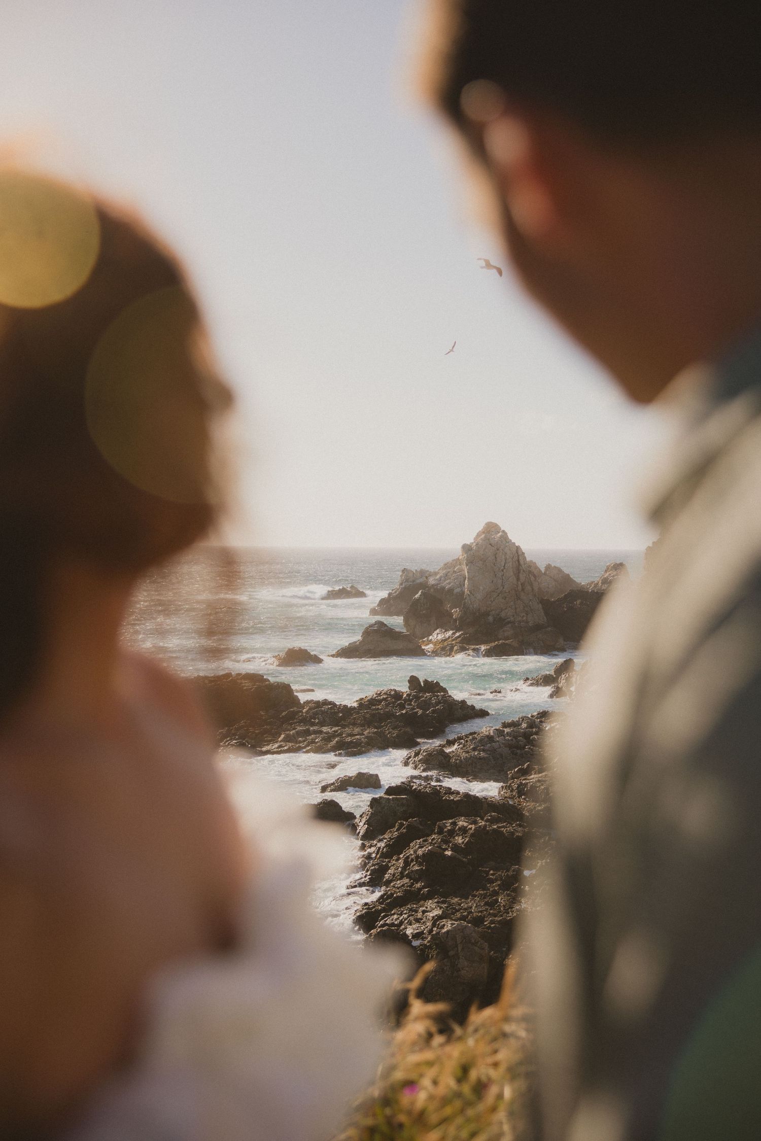 Blurred couple in foreground with dramatic rocky ocean coastline at golden hour in background.