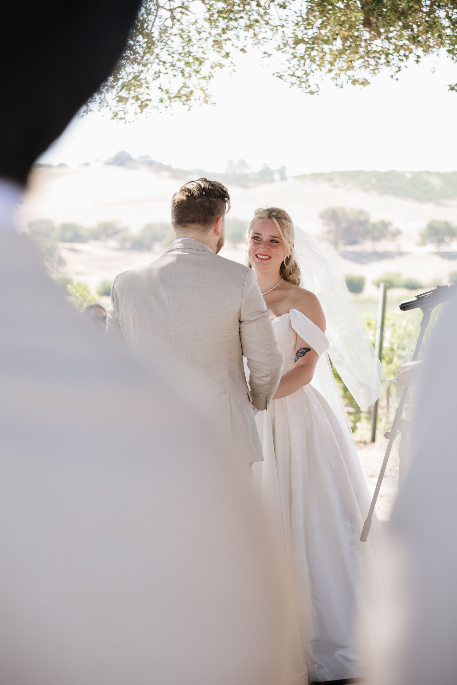 Smiling bride in off-shoulder white gown and veil exchanges vows with groom outdoors at scenic vineyard wedding.