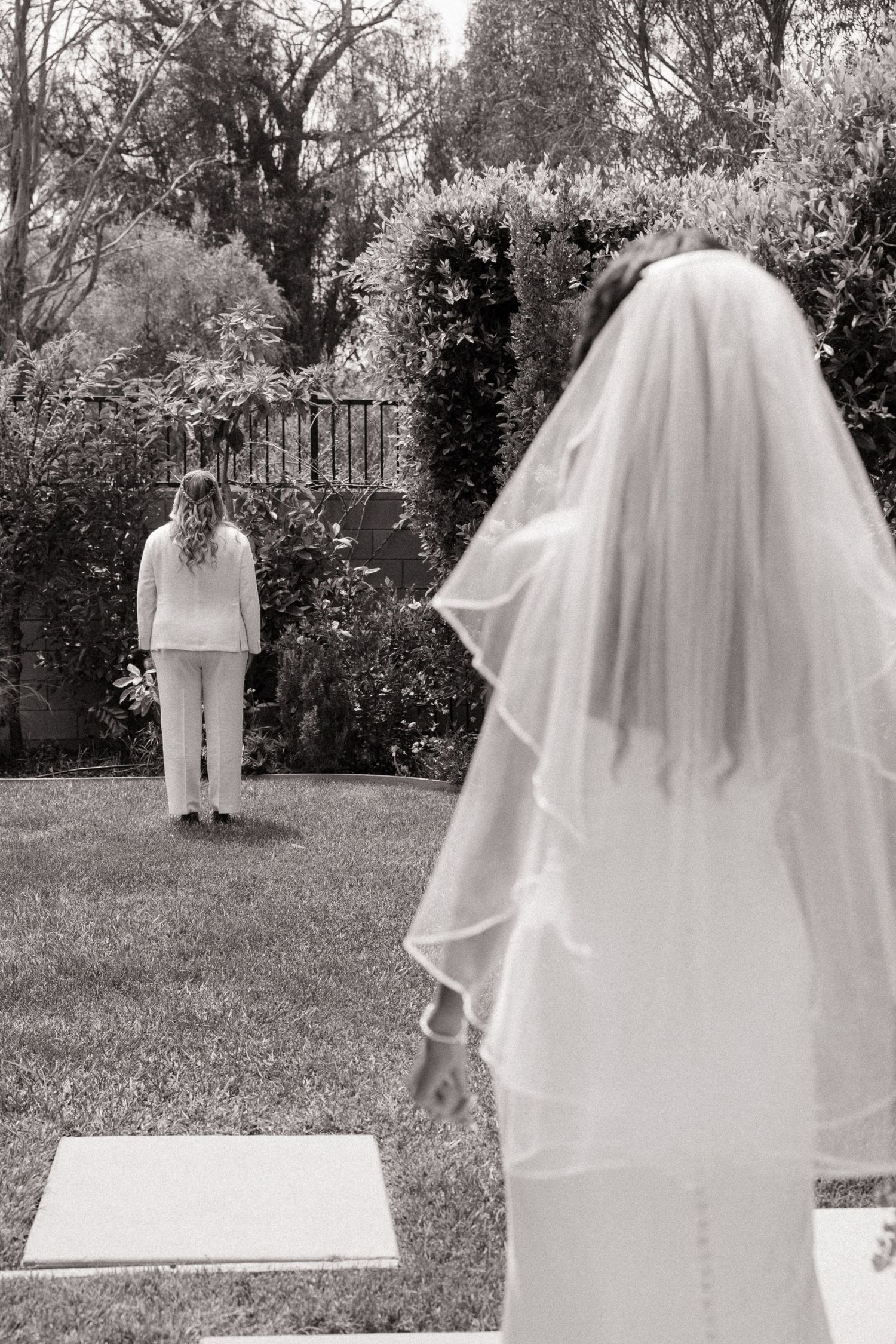 Black and white photo of a bride in a veil approaching her partner in a garden.