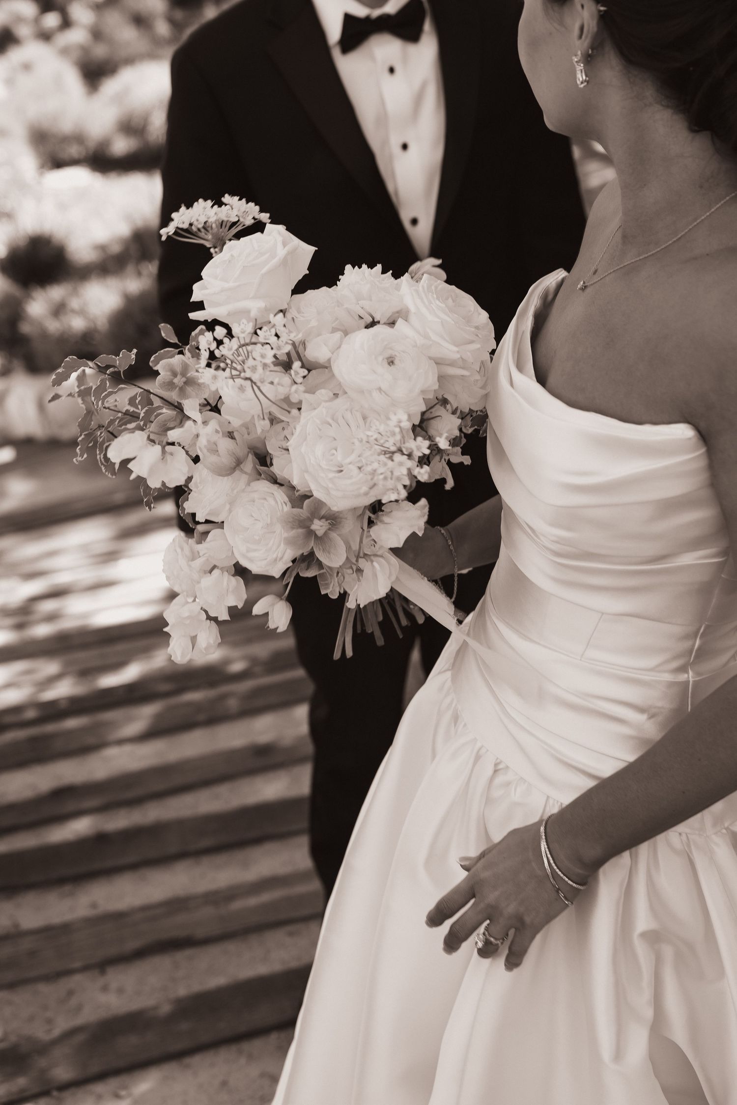 Sepia-toned close-up of bride holding lush white peony and ranunculus bouquet beside groom in black tuxedo.