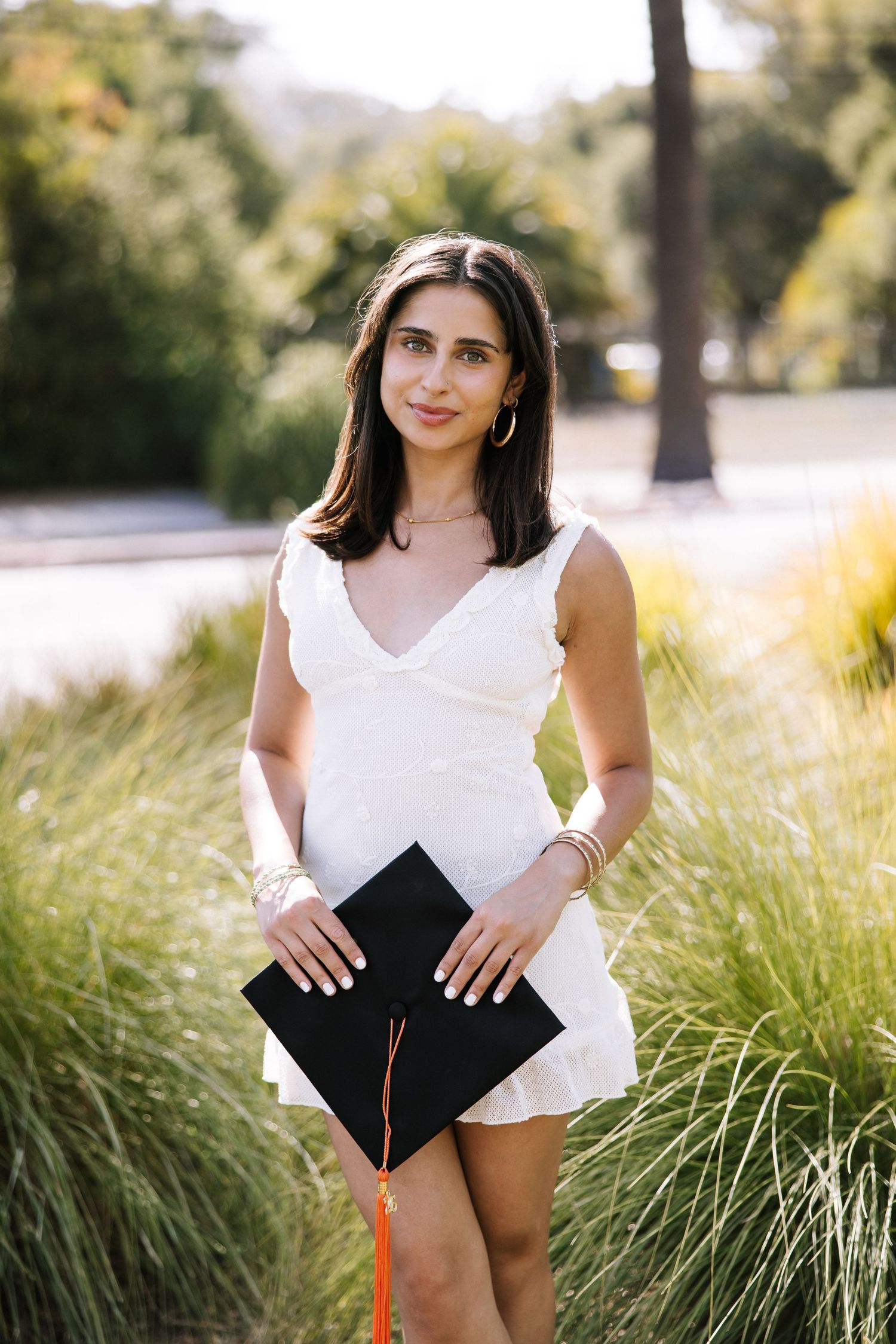 Young woman in white dress holding black graduation cap standing among tall grass outdoors.