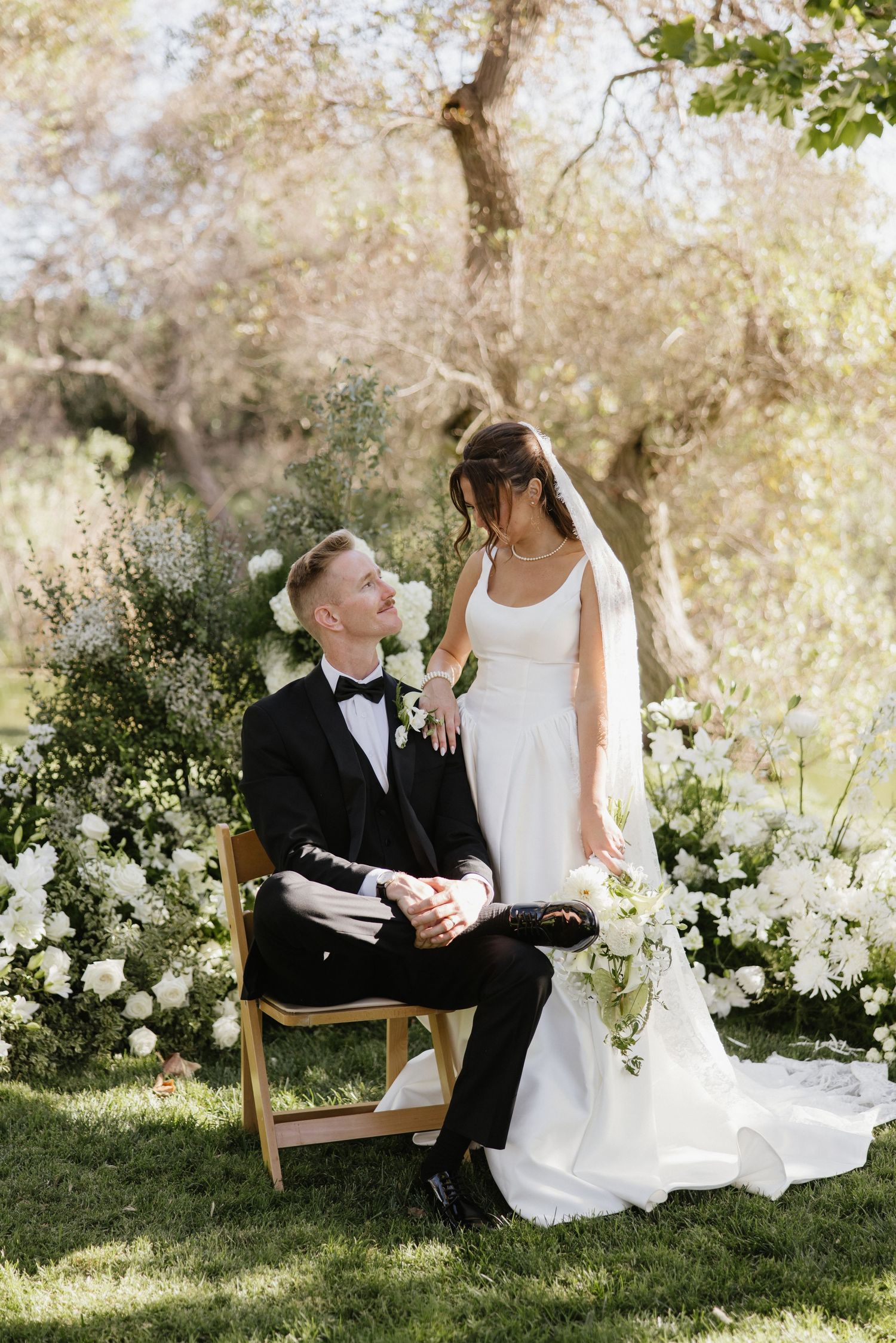 Bride in white gown stands beside groom in black tuxedo seated on wooden chair, surrounded by white florals outdoors.