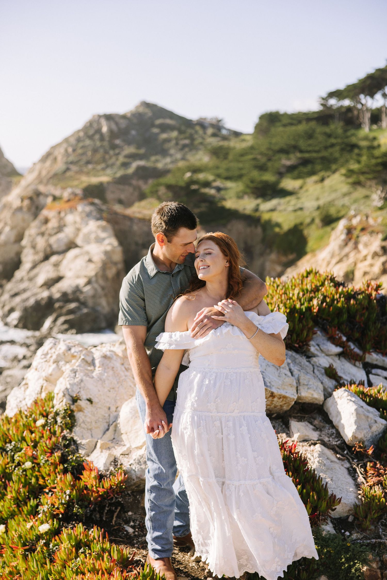 Couple embracing on rocky coastal terrain with cypress trees and colorful ice plants in background.
