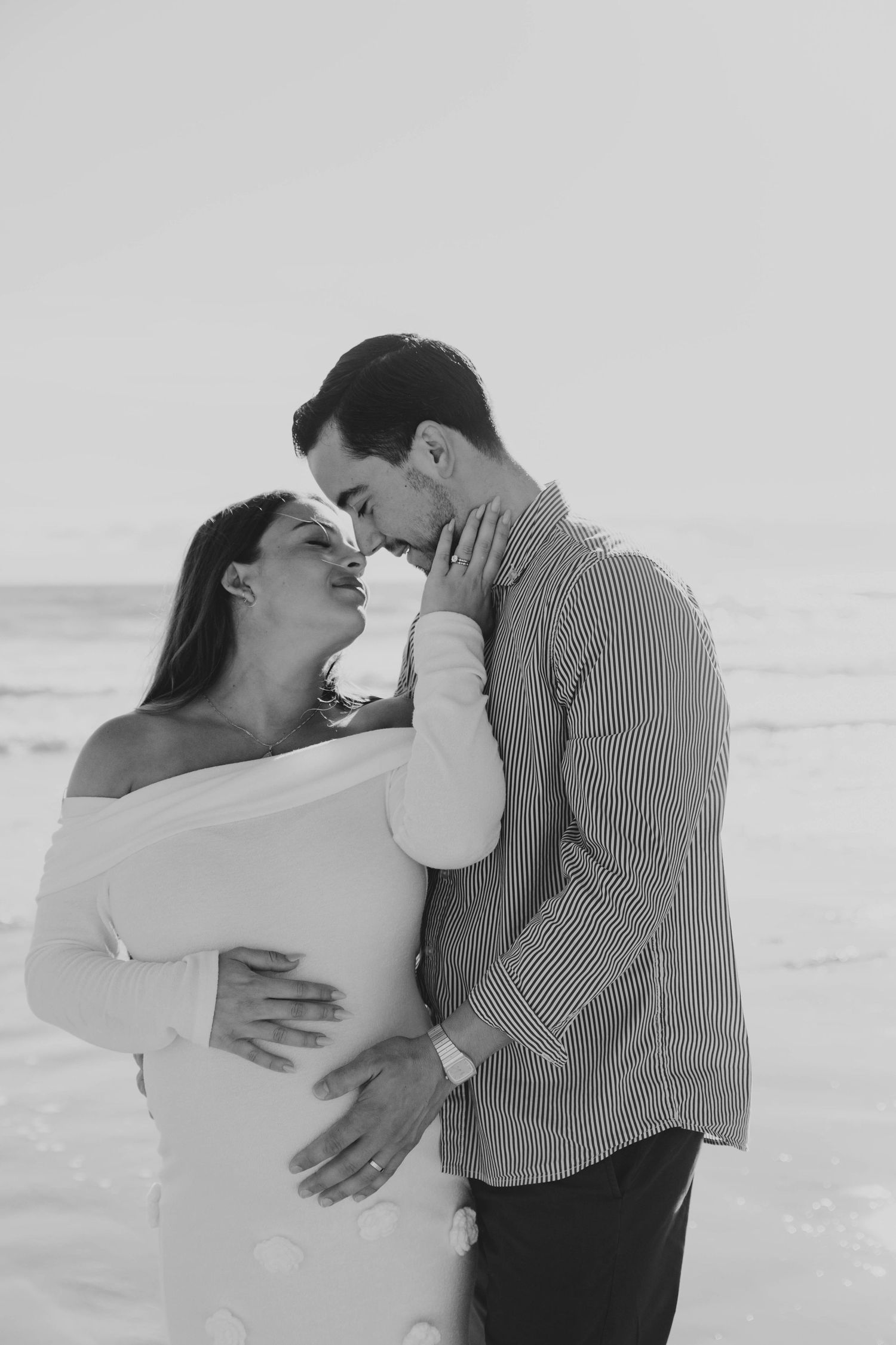Black and white romantic beach photo of pregnant couple almost kissing, man cradling womans baby bump.