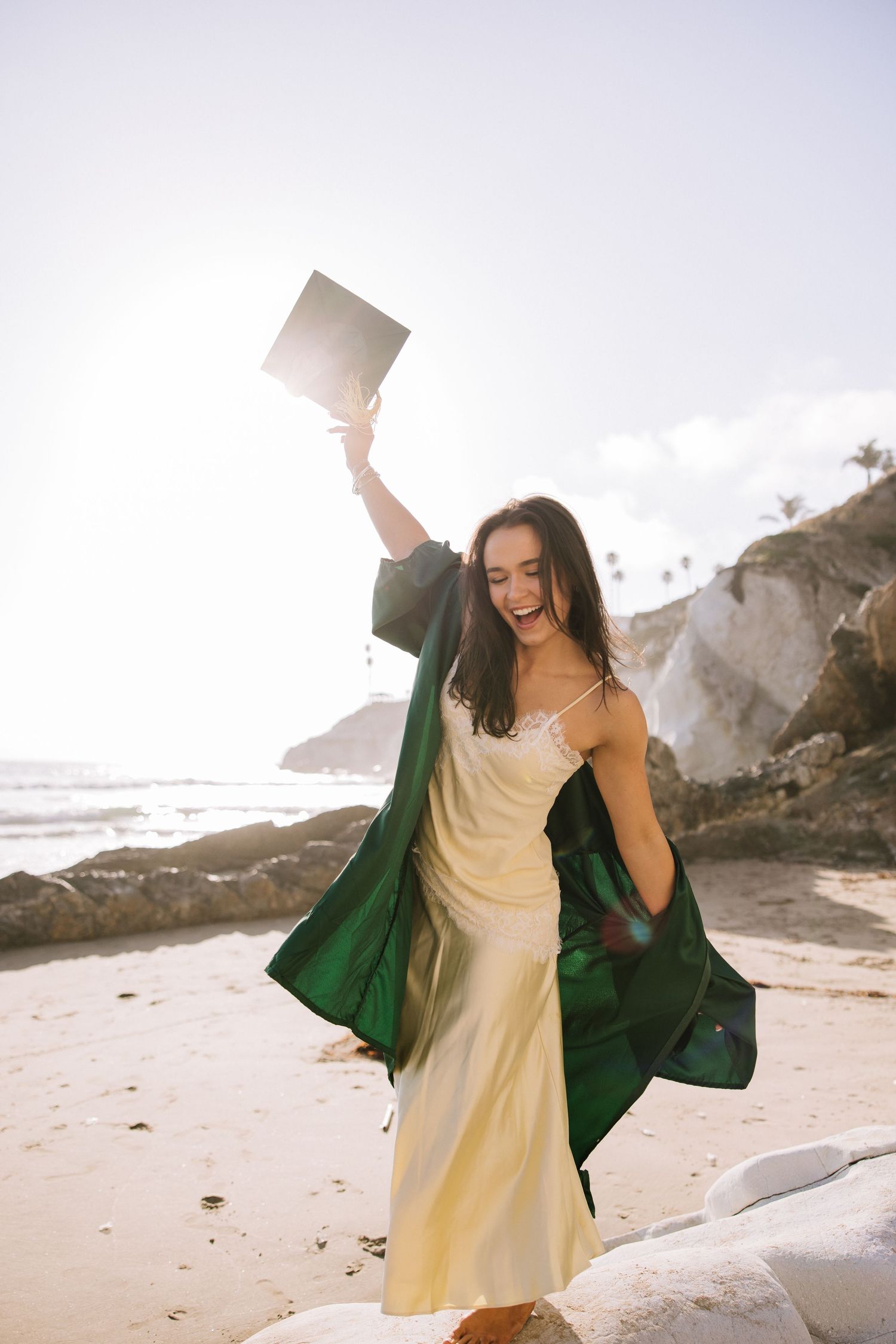 Joyful brunette woman in green graduation gown raising cap on sunny beach for graduation photos.