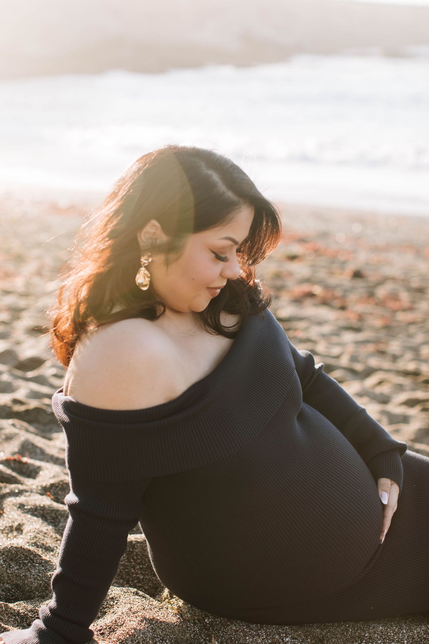 Pregnant woman in black off-shoulder dress sitting on sandy beach in warm sunset light.