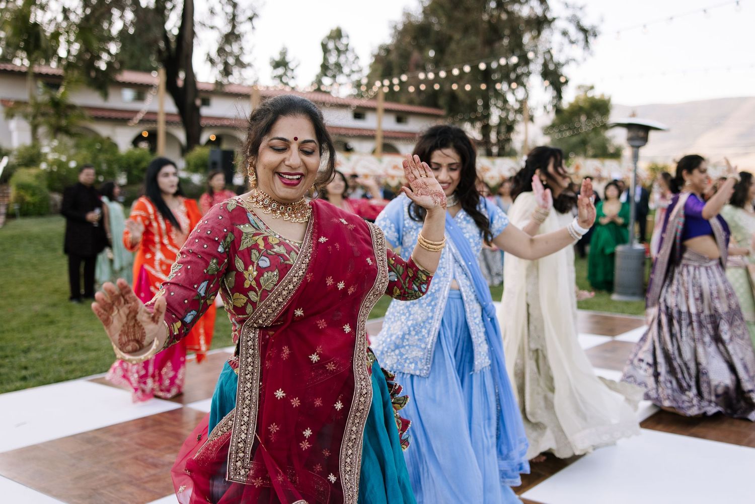Women in vibrant traditional Indian sarees dancing joyfully at an outdoor wedding celebration.