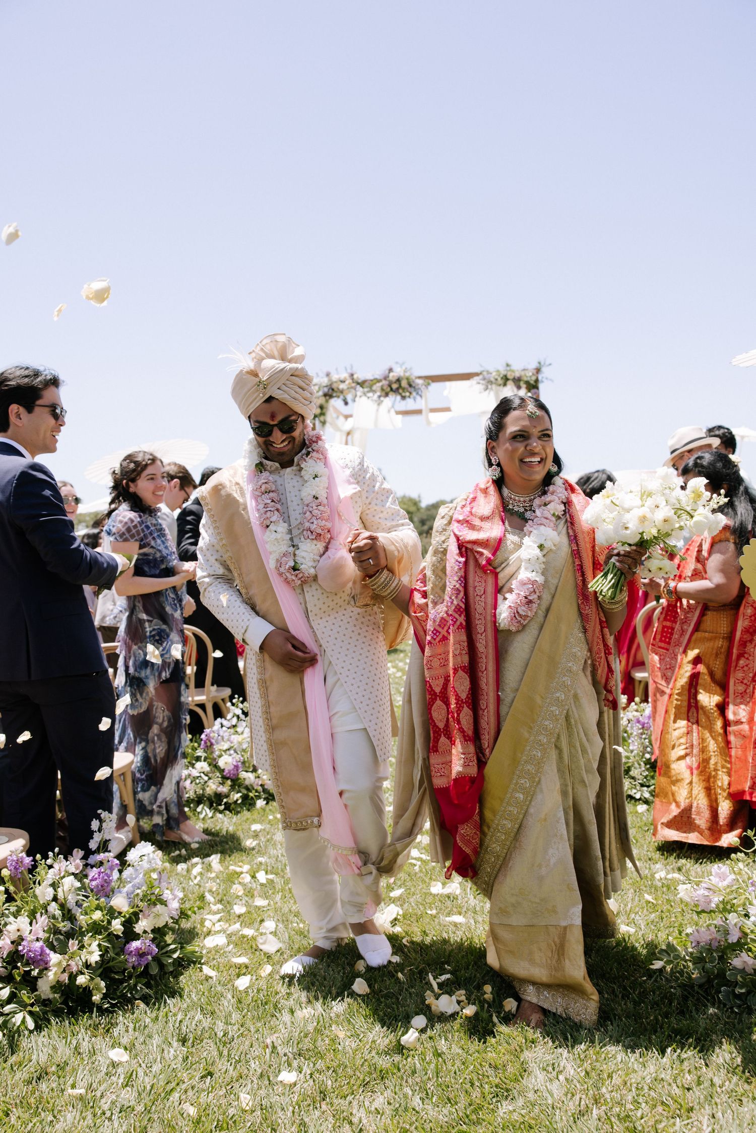 Indian groom in white sherwani and turban walks with mother in red saree after outdoor wedding ceremony.