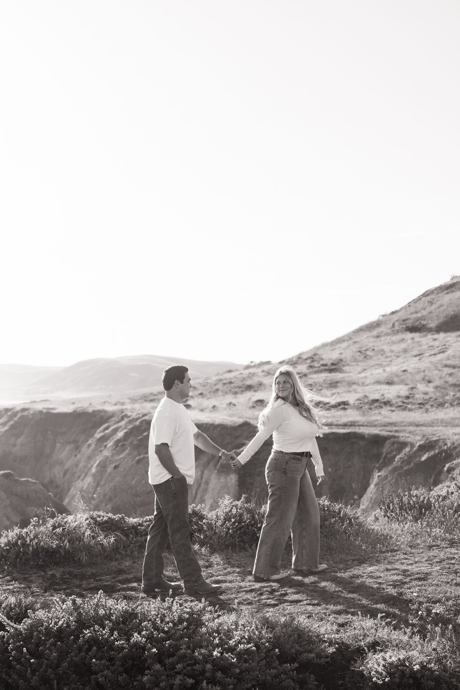 Couple holds hands walking on coastal cliffs in black and white engagement photo.
