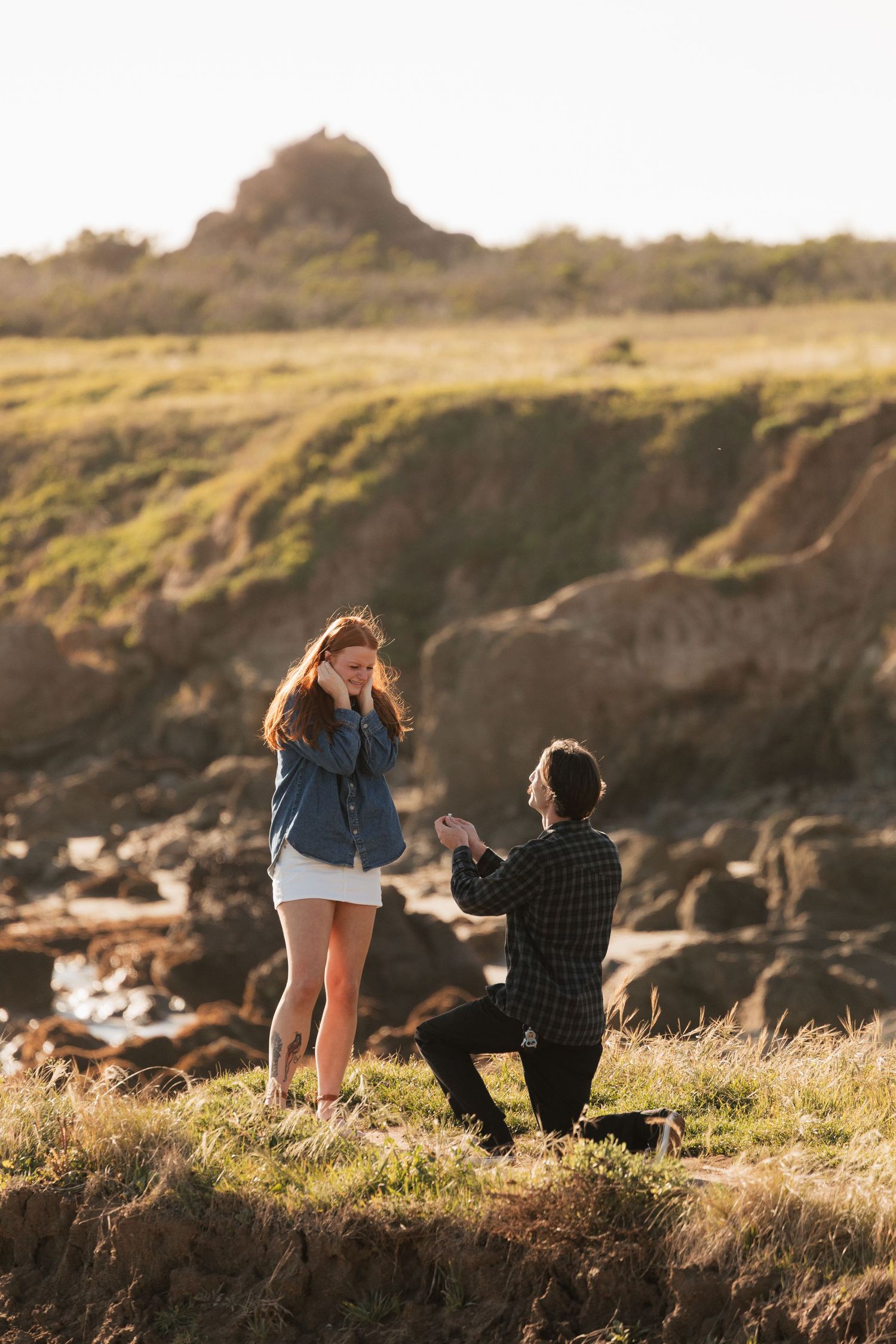 Man kneeling on coastal cliff proposing to surprised woman in denim jacket and white skirt at golden hour.