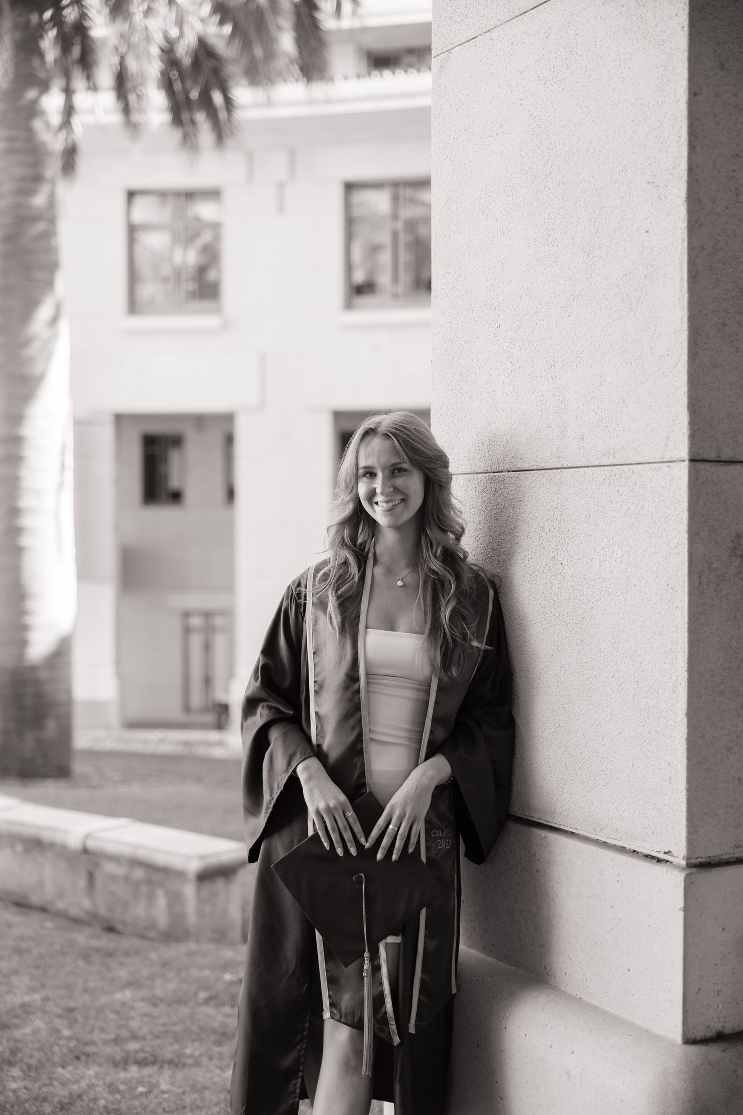 Smiling graduate in black gown leans against stone wall holding cap outdoors.