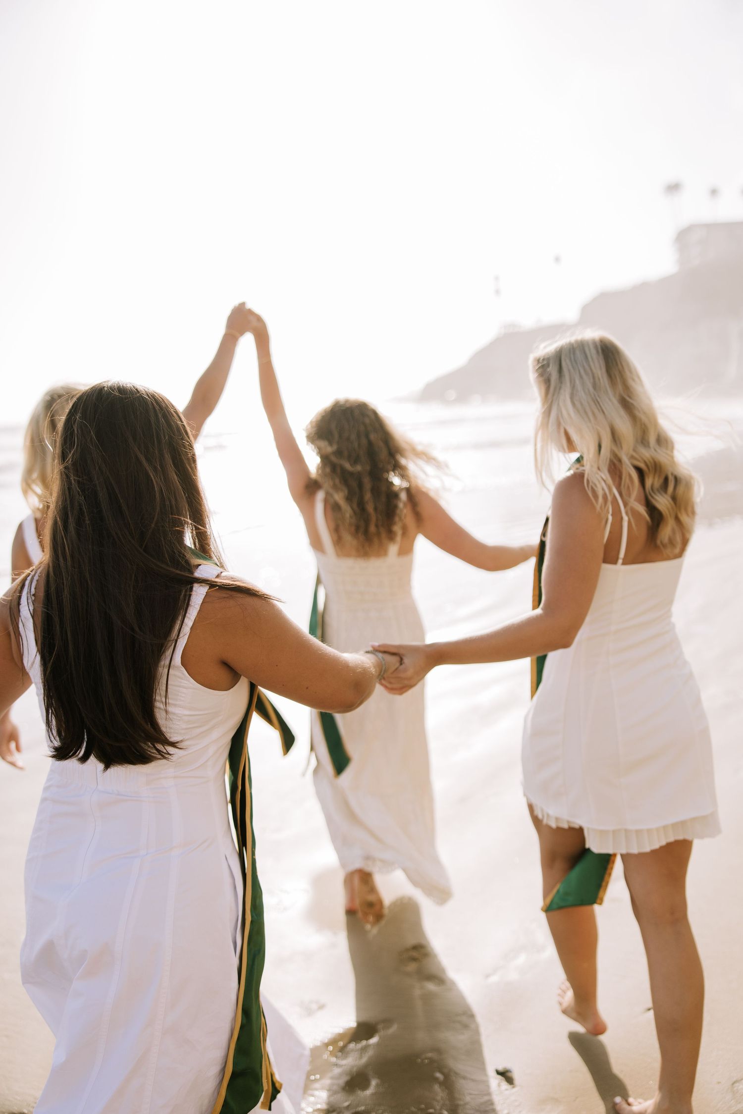 Four female graduates in white dresses and green sashes hold hands walking toward ocean waves.