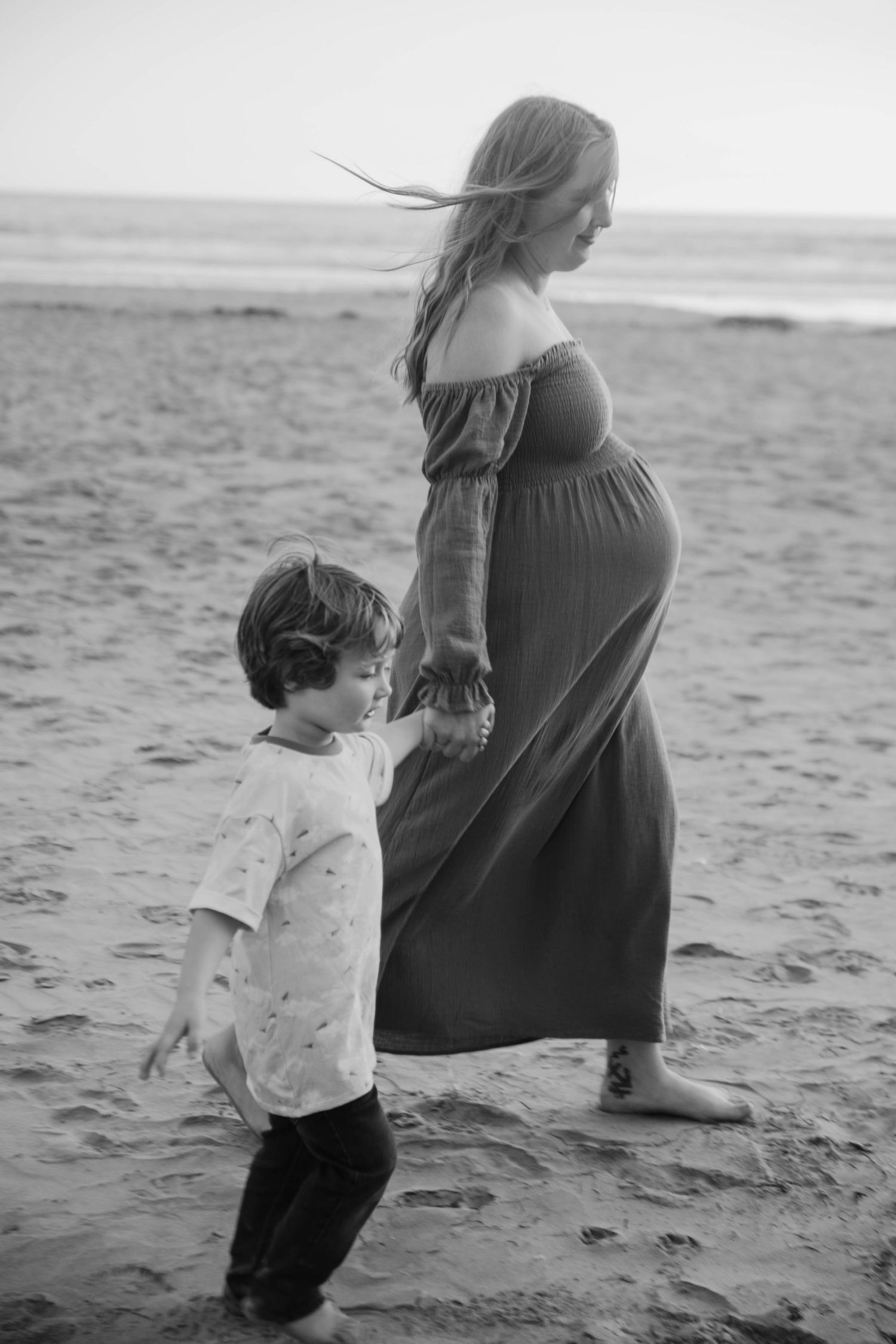 Black and white beach maternity photo of pregnant woman in long dress with young son.