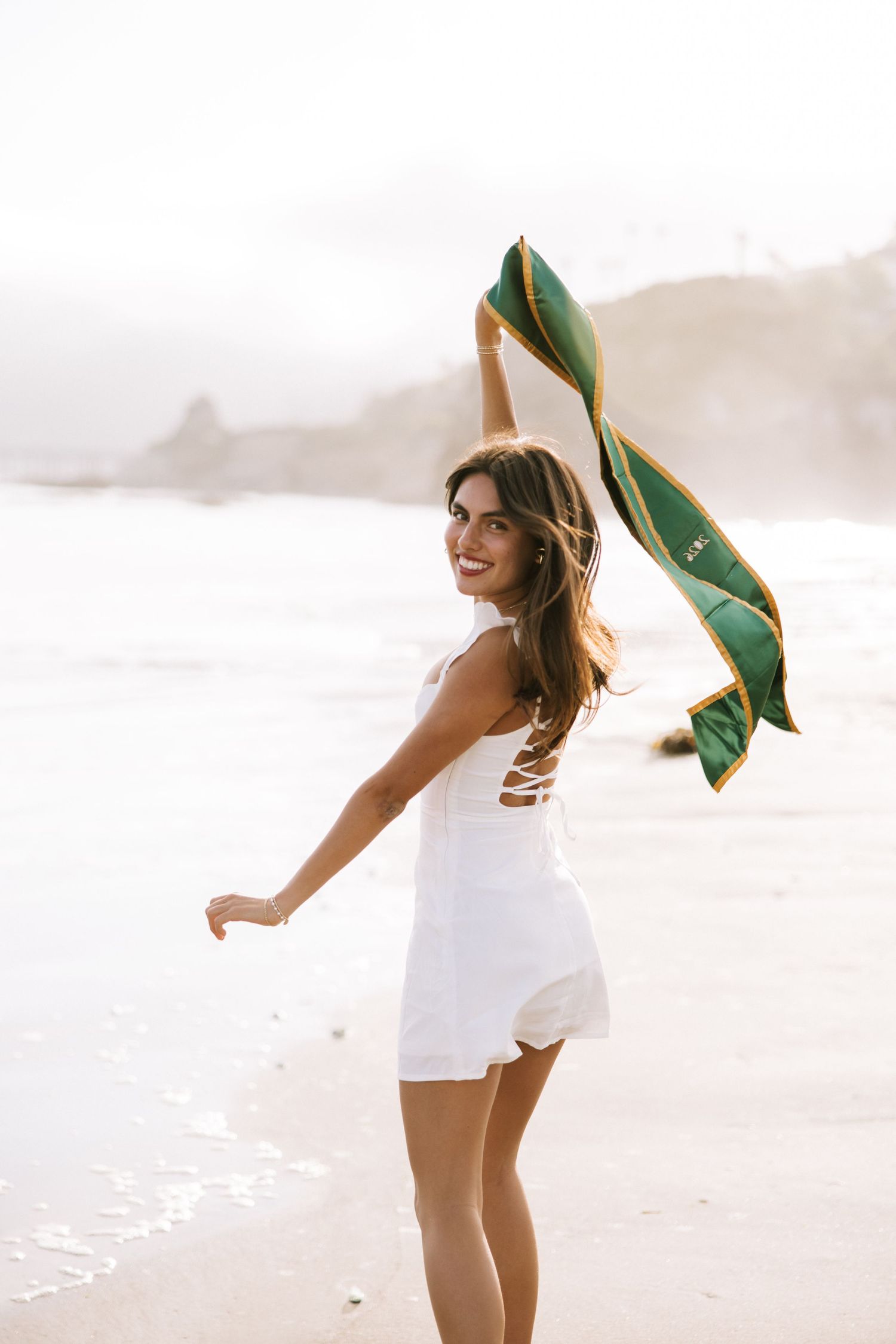 Joyful woman in white romper waving a green graduation stole on a sunny beach shoreline.