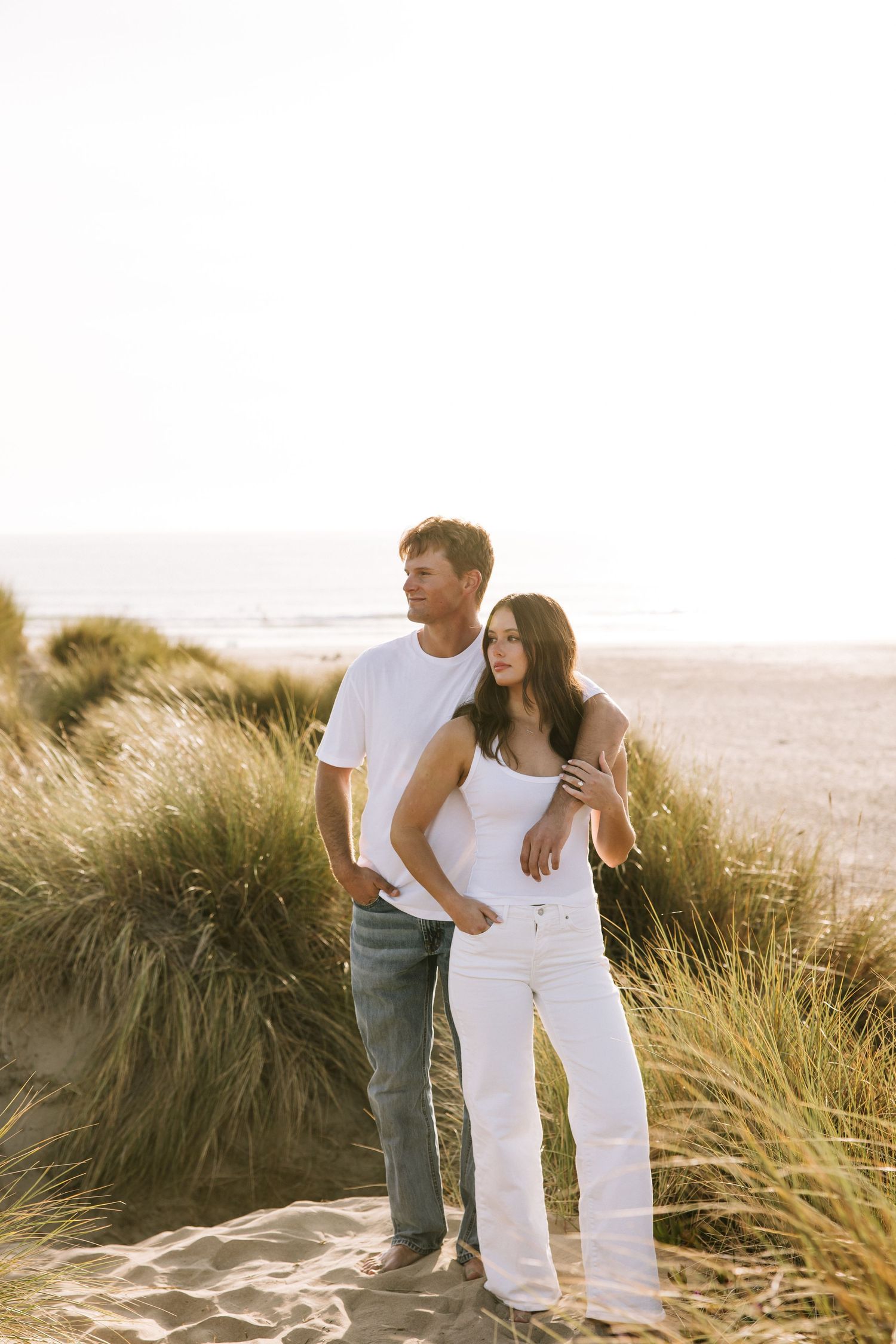 Couple embracing in golden beach grass dunes at sunset with ocean in background.
