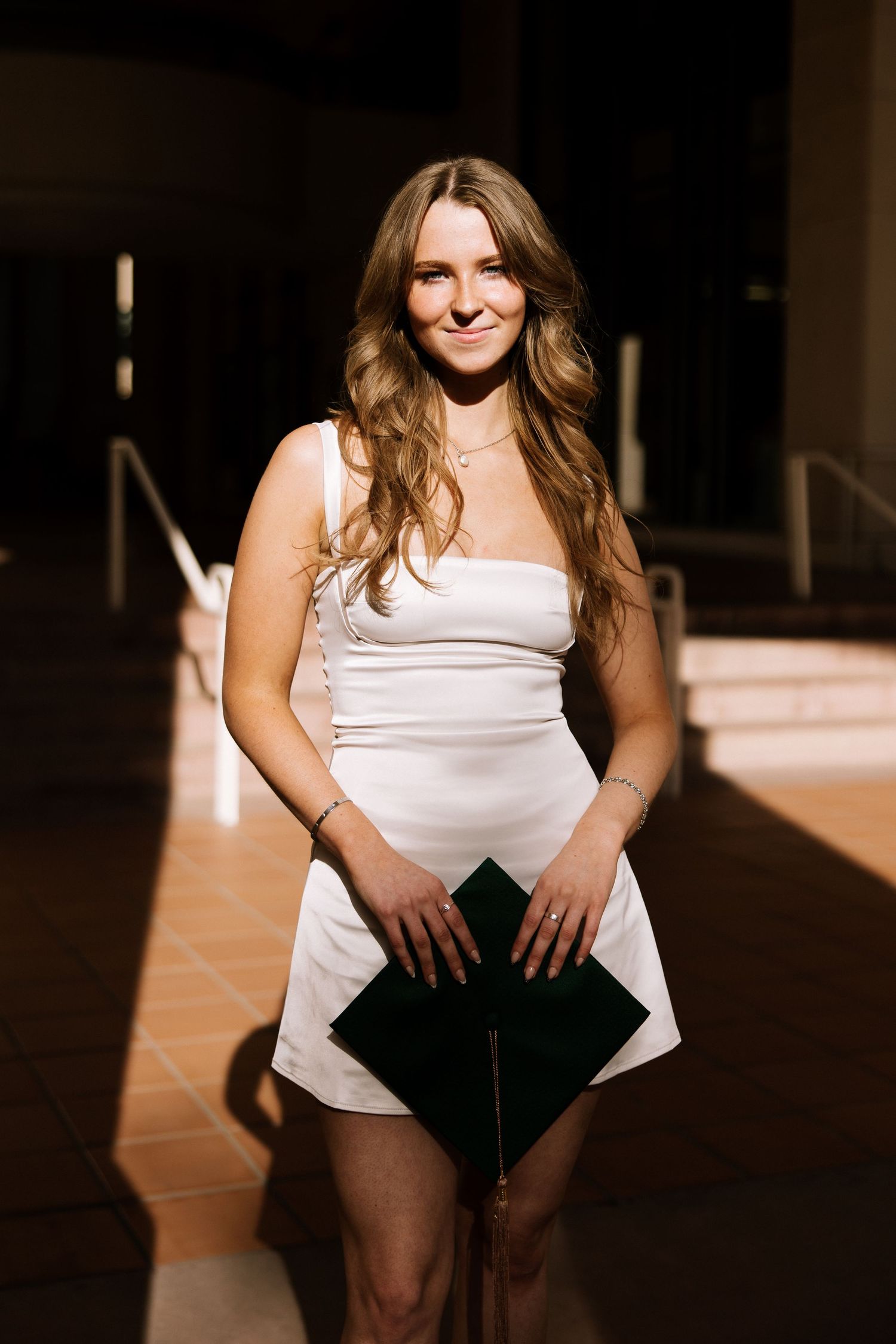 Young woman in white mini dress holds graduation cap in dramatic architectural lighting.