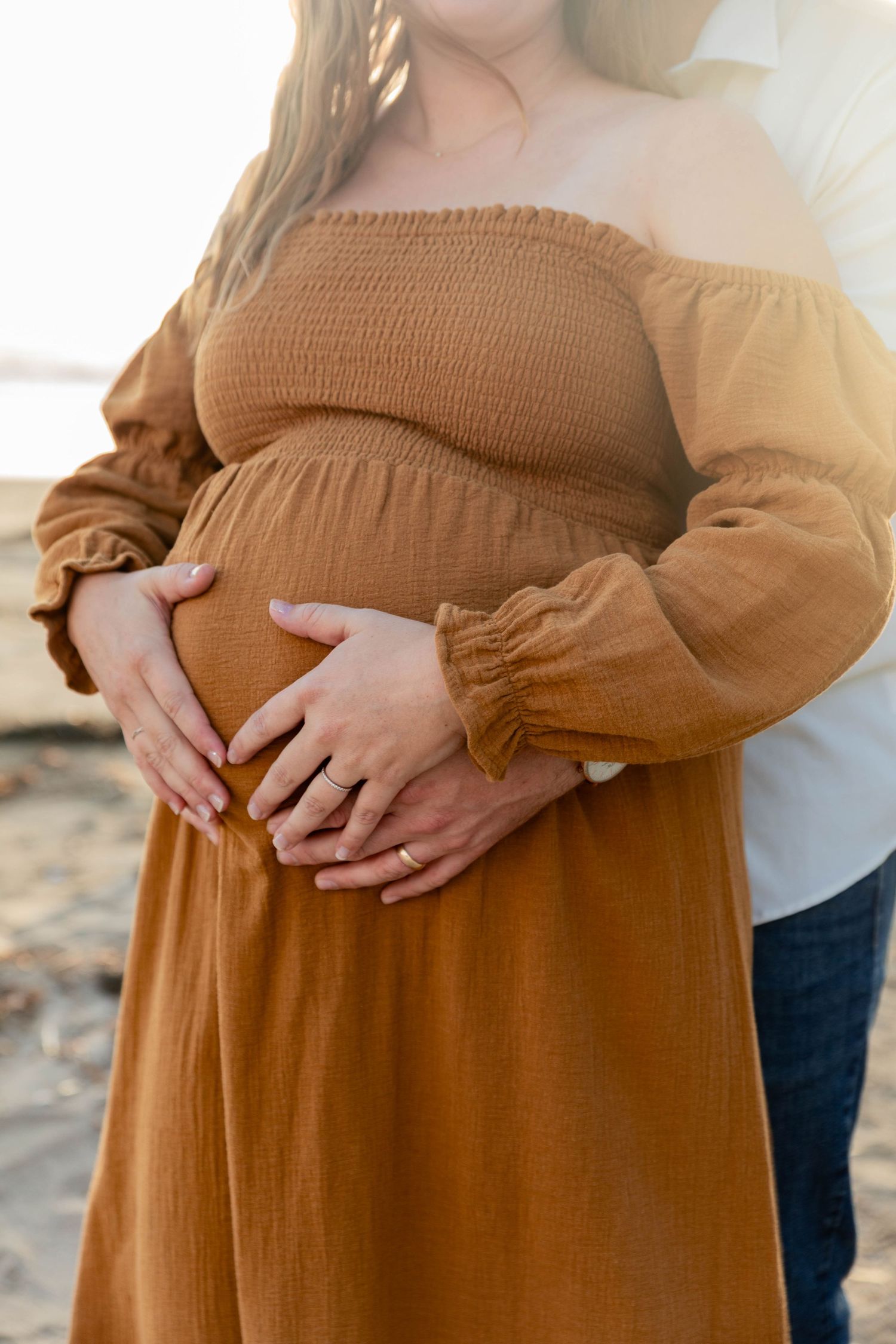 Couple holding pregnant belly together, woman in rust off-shoulder dress at beach.