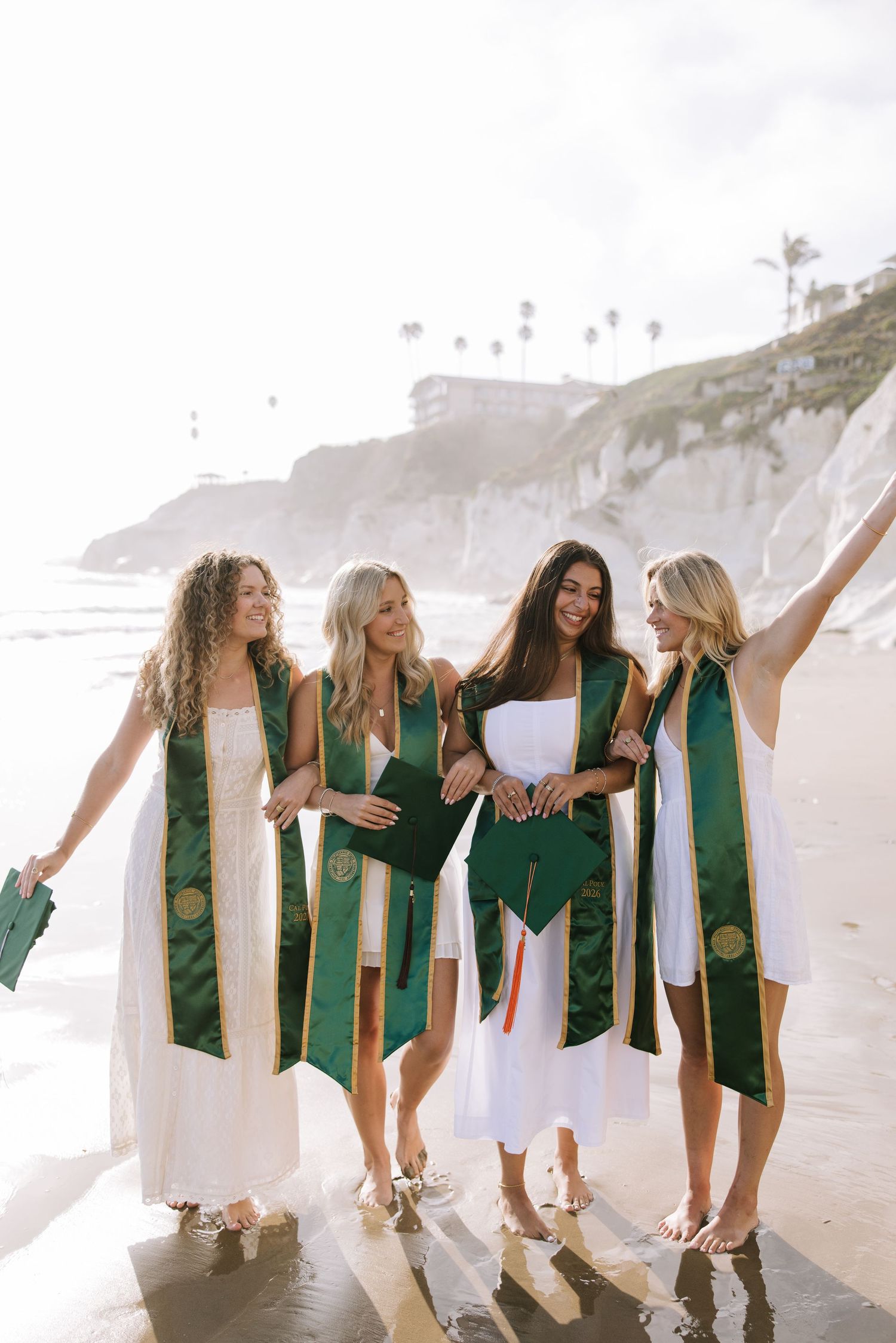 Four happy female graduates in green sashes walk along sunny beach holding graduation caps.