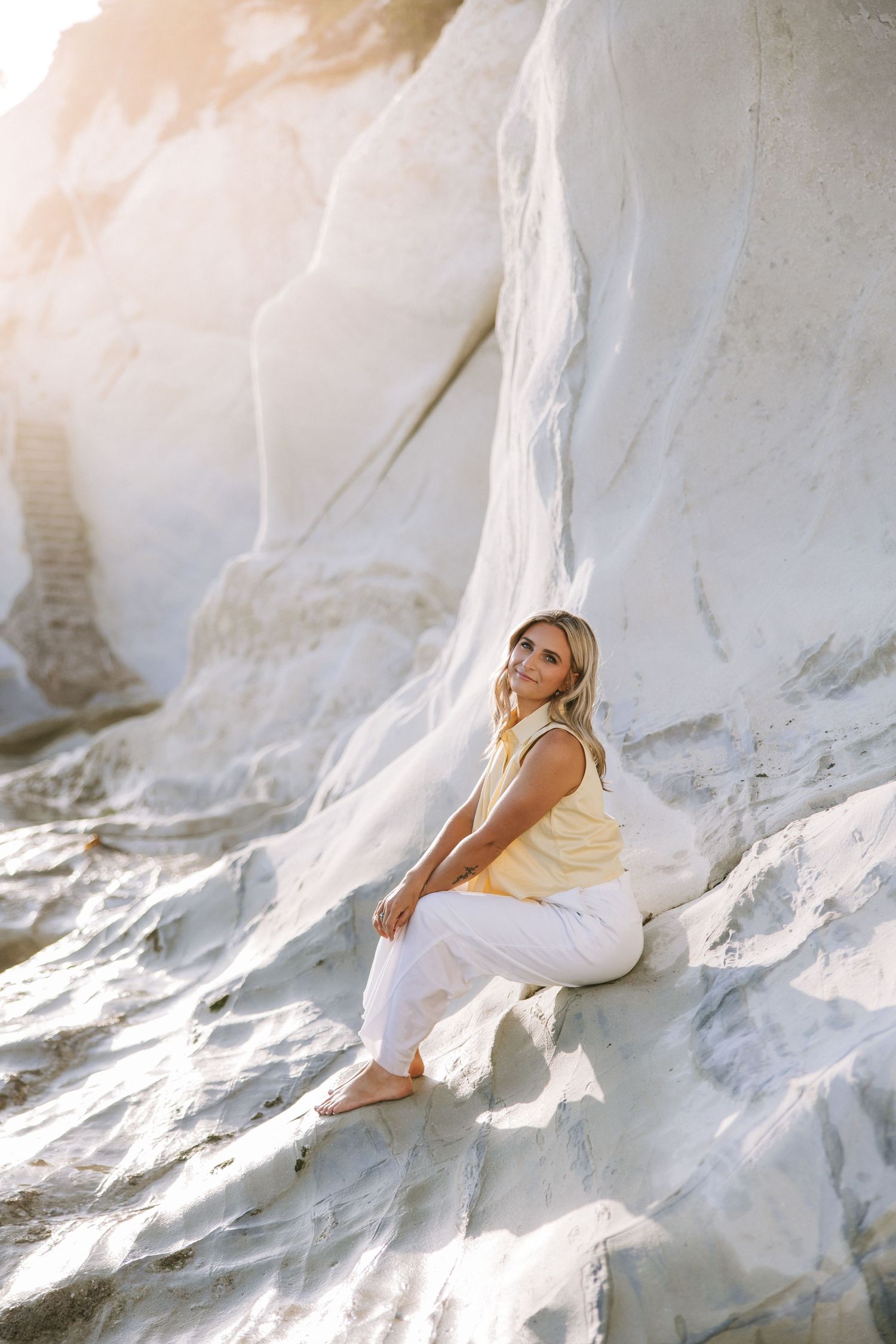 Barefoot blonde woman in yellow top and white pants sitting on white rock cliff in sunlight.