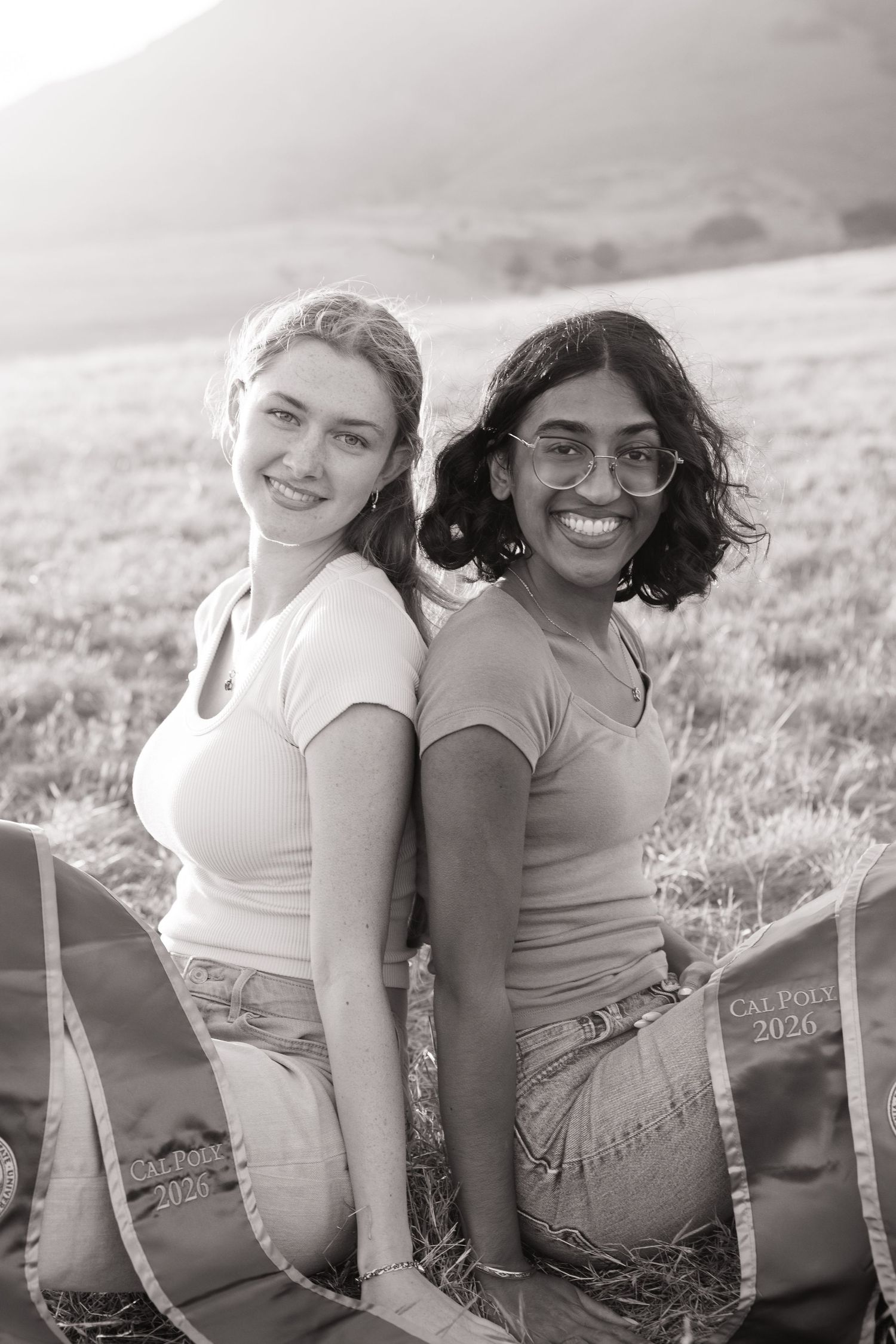 Black and white photo of two smiling women sitting back-to-back in field with Cal Poly 2026 sashes.
