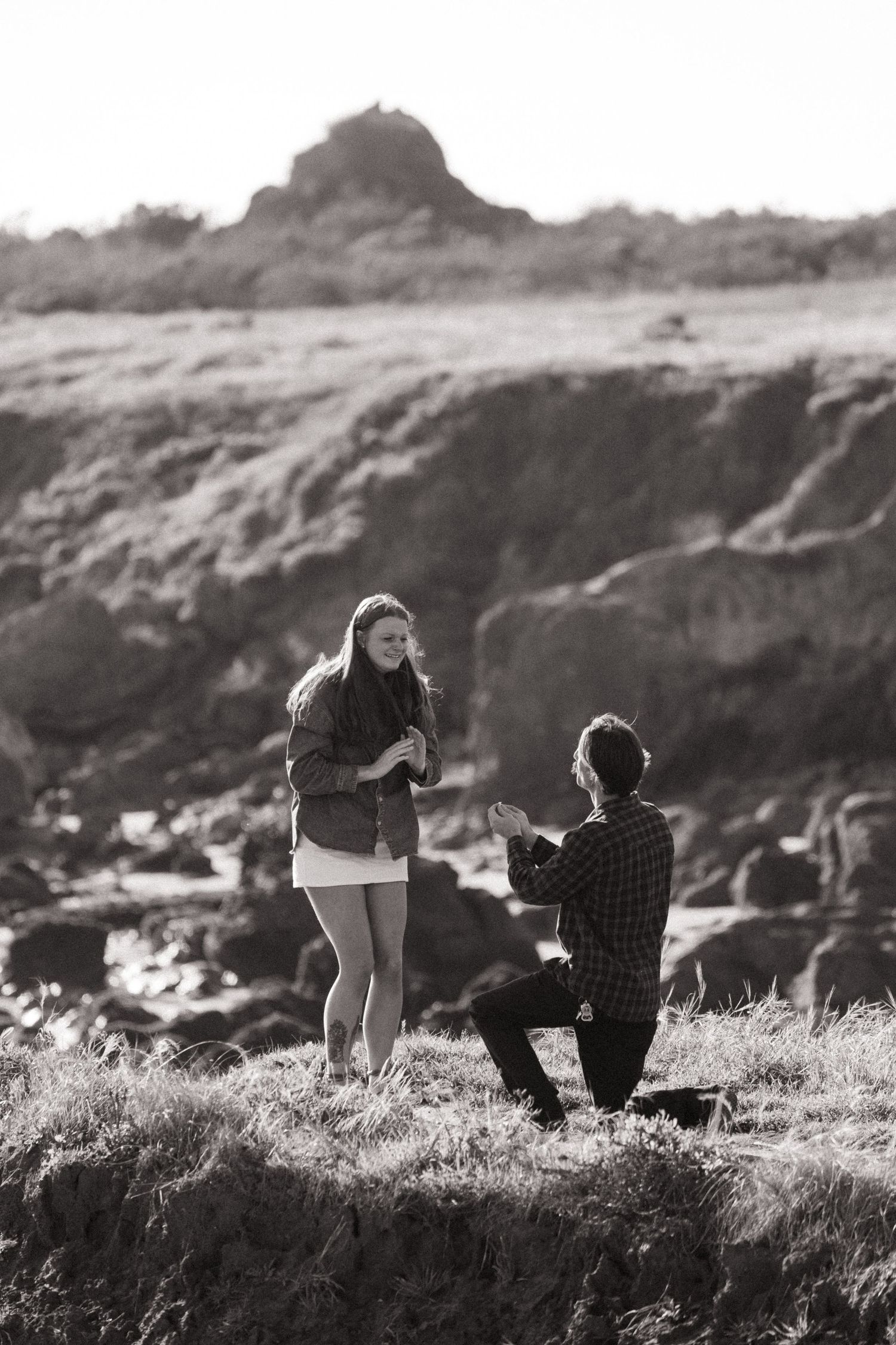Black and white photo of a man proposing on one knee to a woman on rocky coastal terrain.
