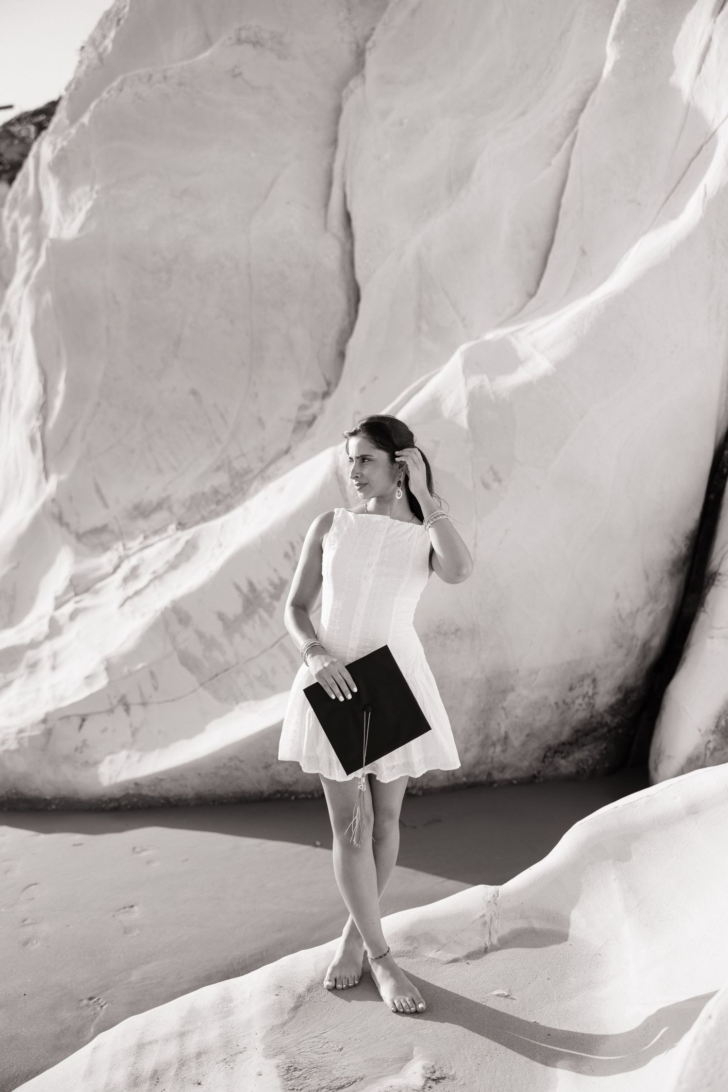 Black and white portrait of woman in white dress holding diploma beside large white rocks.