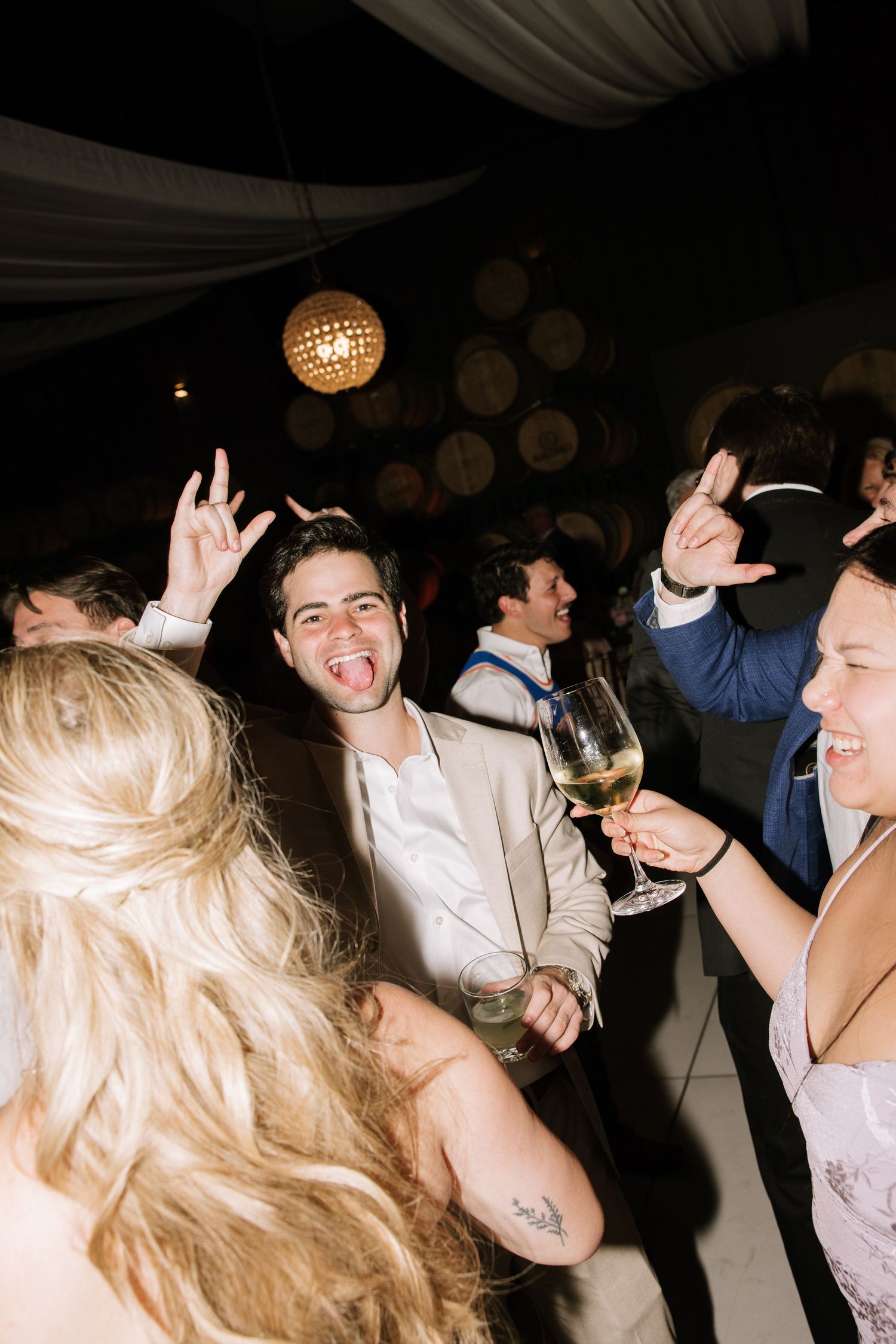 Wedding guest in tan suit makes rock sign with tongue out while dancing at lively reception party.