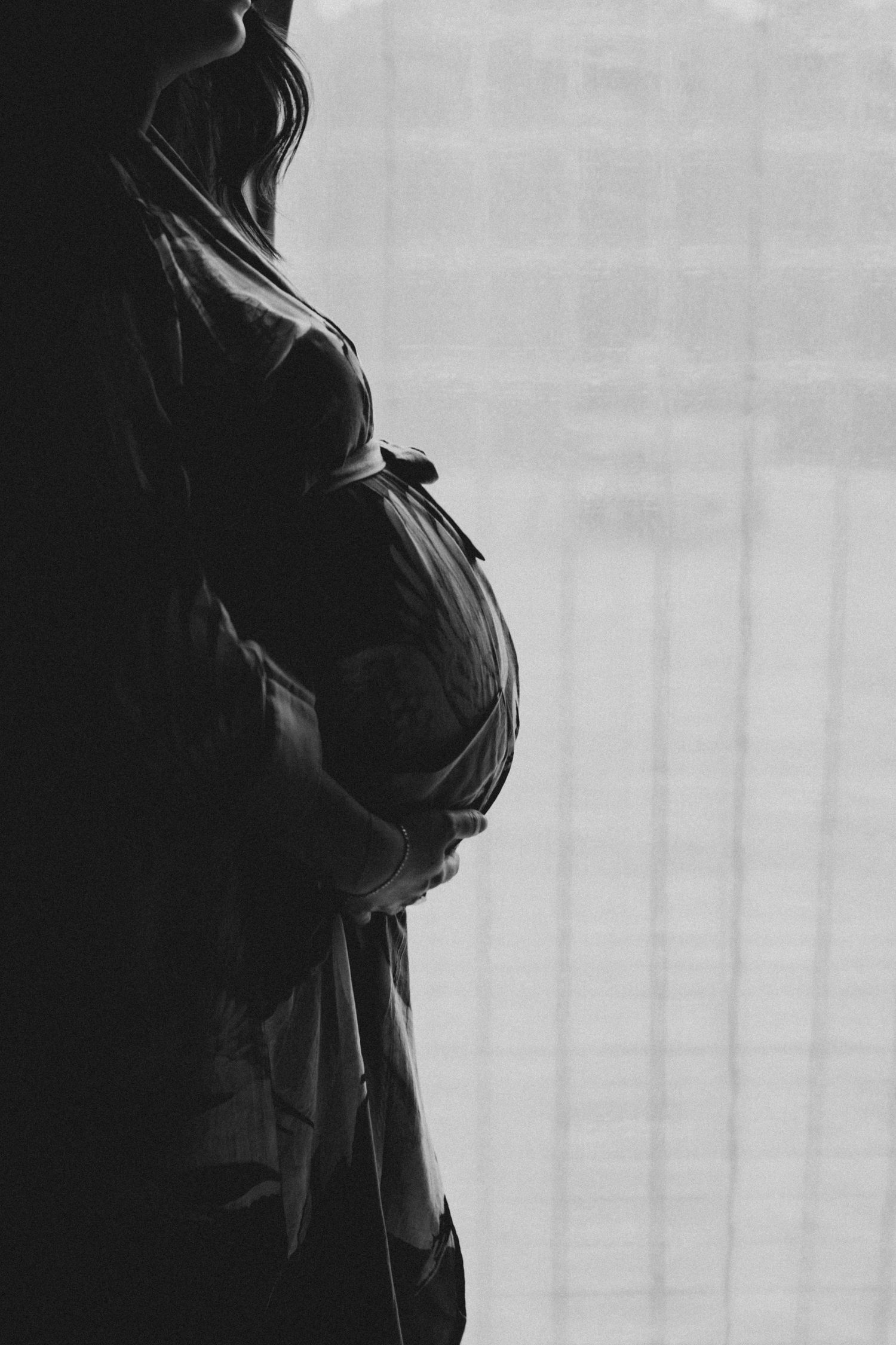 Black and white silhouette of a pregnant woman cradling her baby bump while standing near a window.