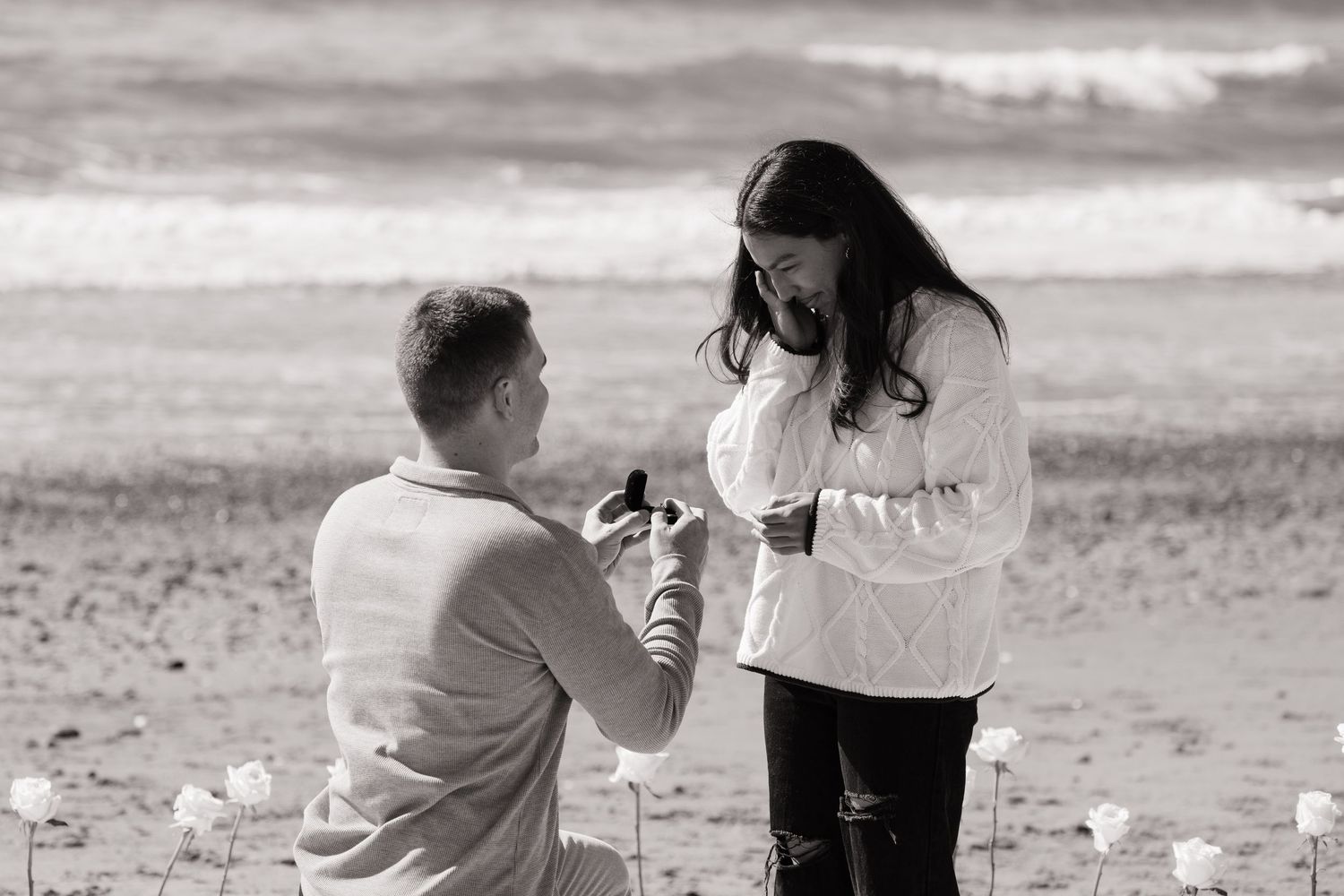 Black and white photo of a man proposing on a beach surrounded by white roses.