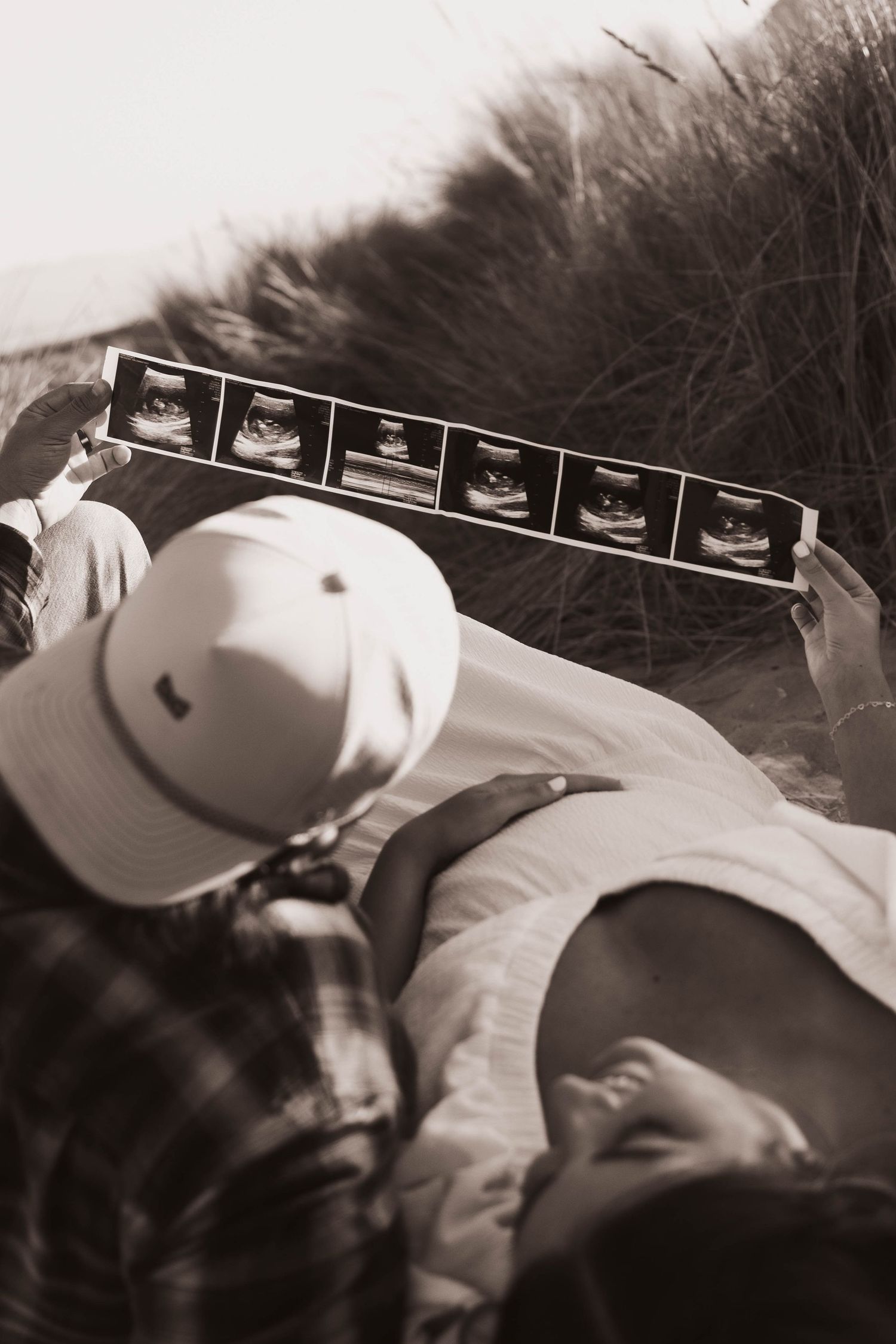 Overhead sepia-toned photo of couple holding ultrasound strip while sitting in beach grass dunes.
