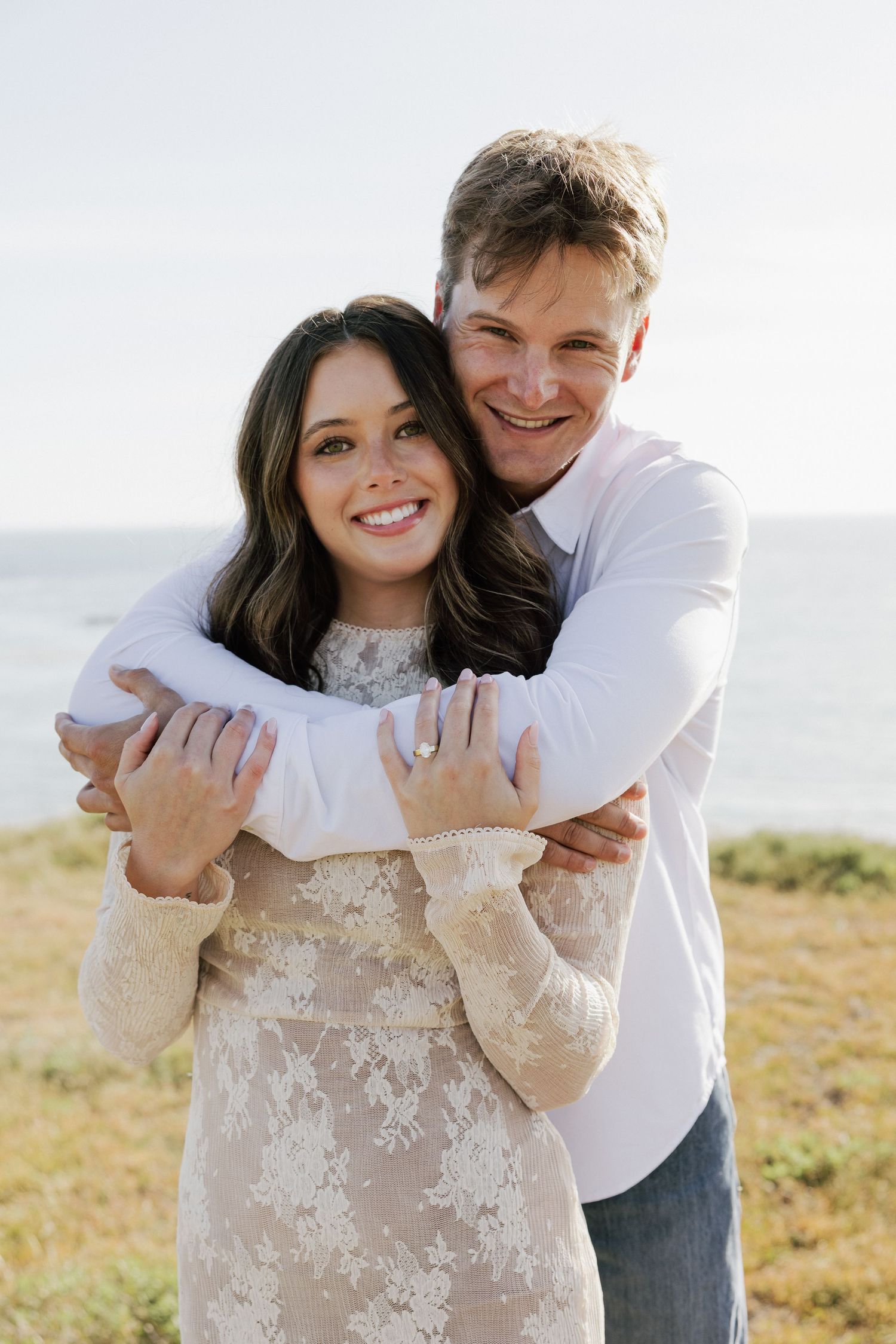 Happy couple embracing on coastal hillside, woman in lace dress showing engagement ring near ocean.