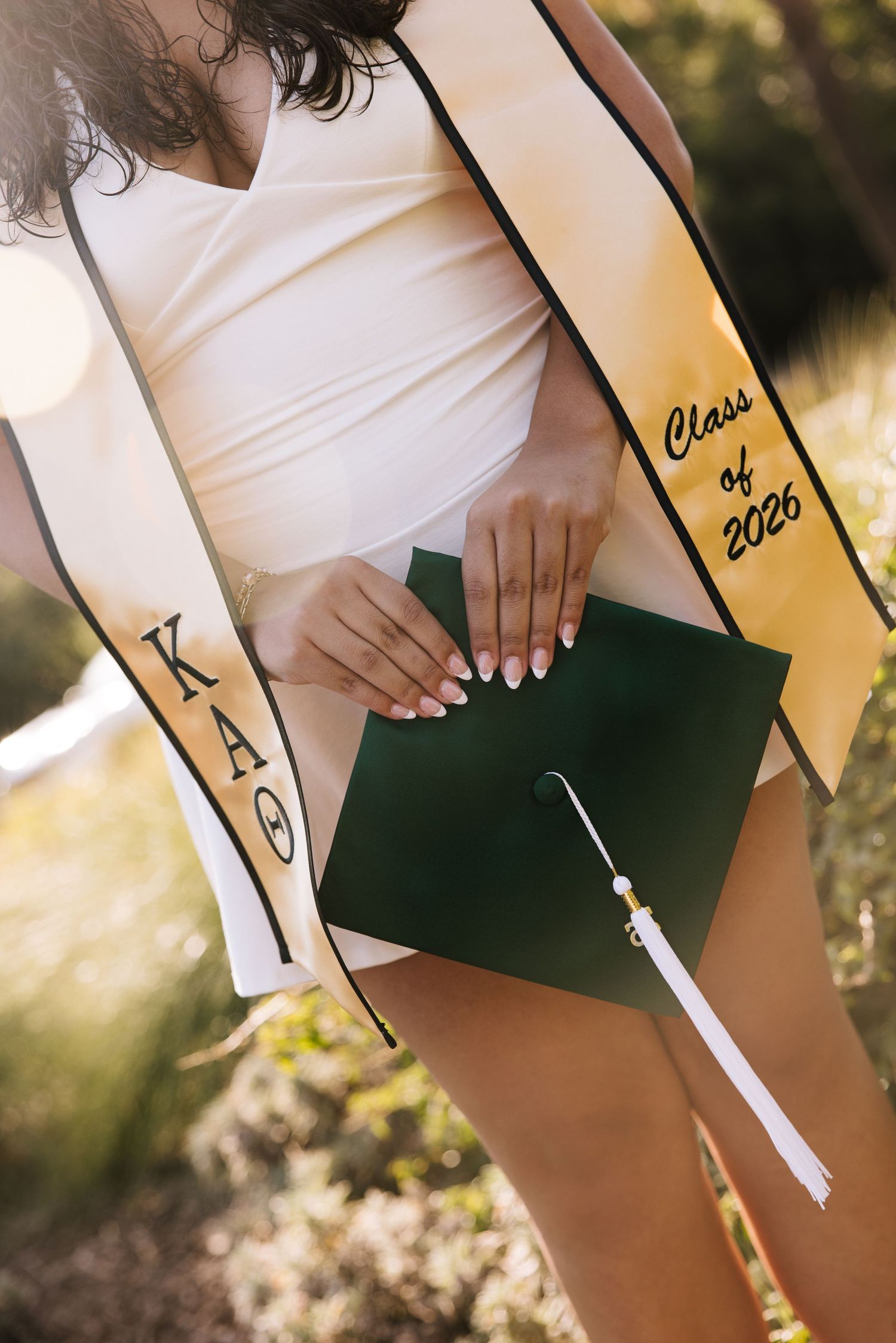Graduate holds green cap wearing Class of 2026 stole and KAO sorority sash outdoors.