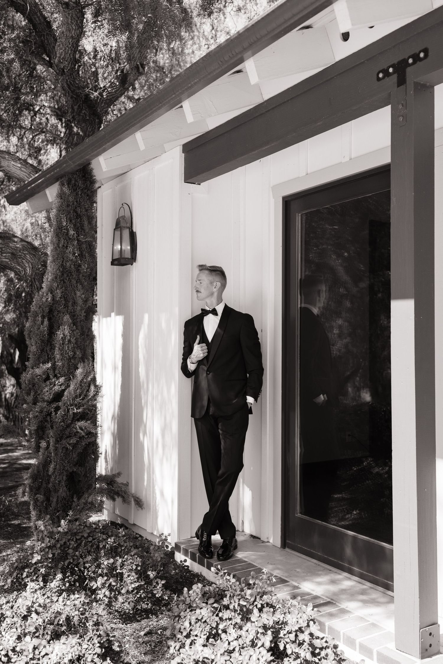 A groom in a black tuxedo and bow tie leans against a white building exterior, looking up thoughtfully.