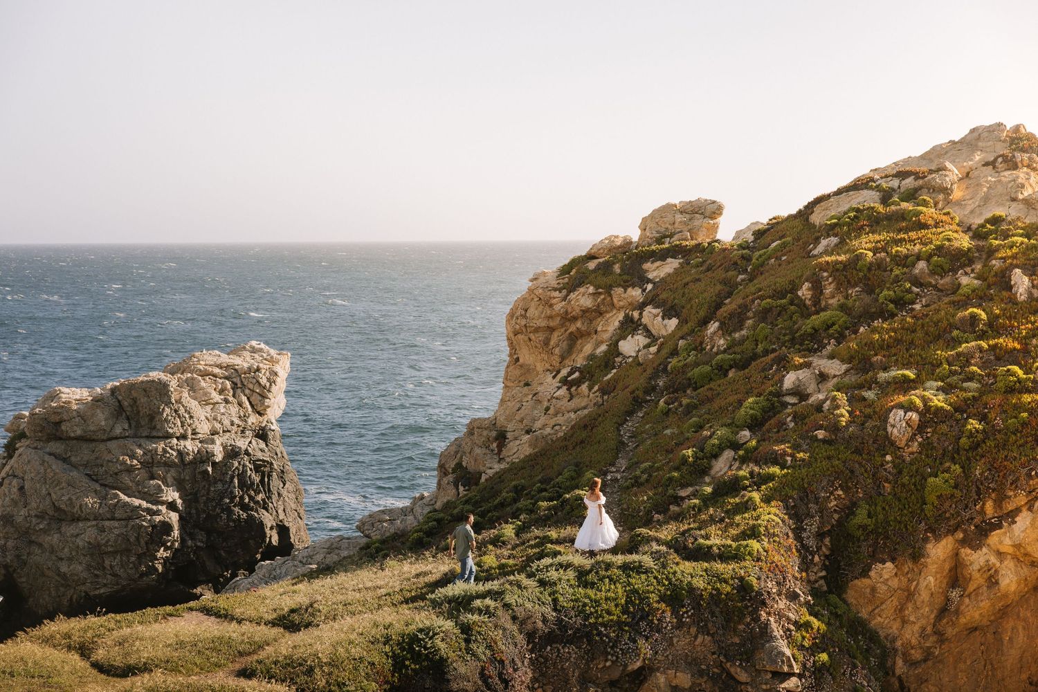 Bride and groom stand on dramatic coastal cliffs overlooking the deep blue ocean at sunset.