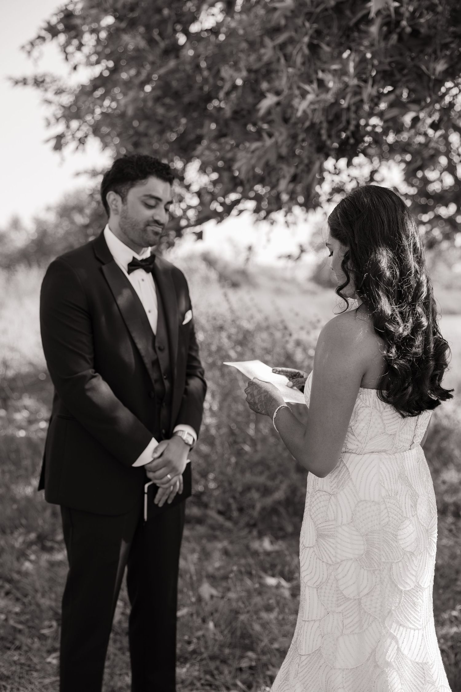 Groom in black tuxedo listening emotionally as bride reads wedding vows in black and white photo.