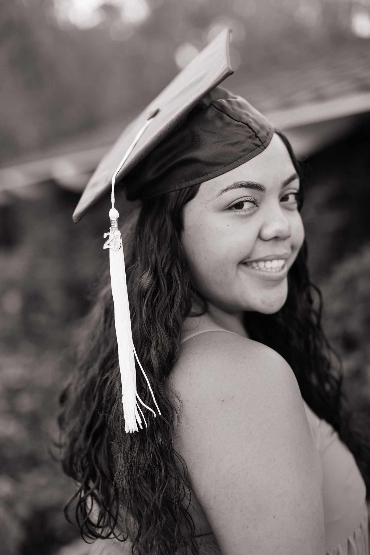 Smiling young woman in graduation cap with tassel poses for black and white senior portrait photo.