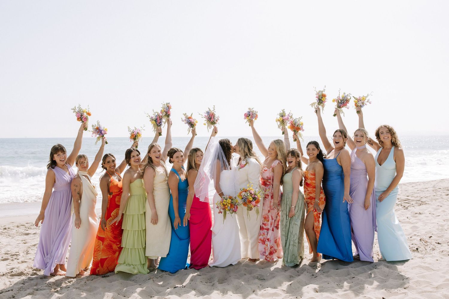 Bridesmaids in colorful dresses raising bouquets with bride on sandy beach.