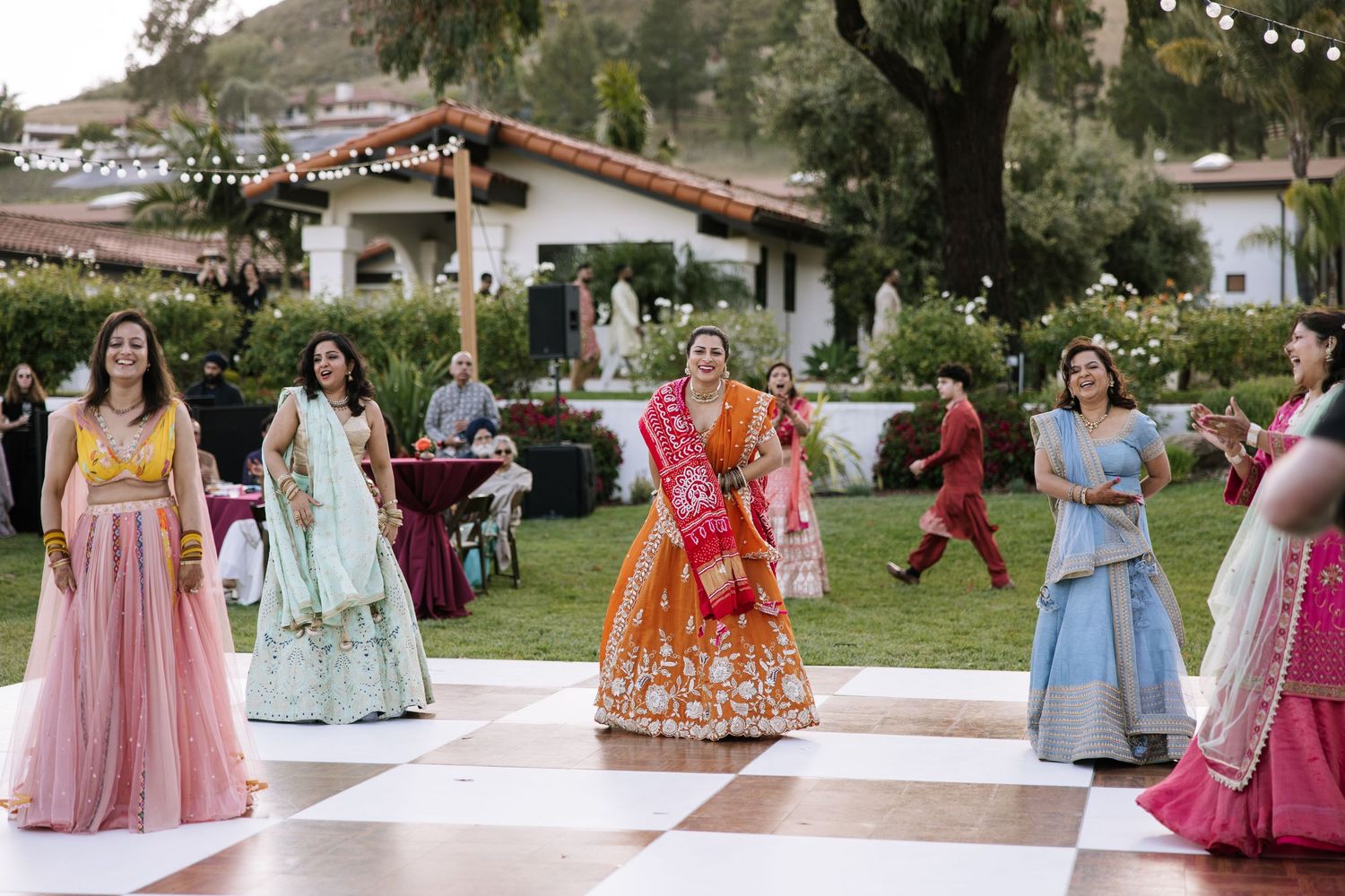 Women in colorful lehengas dance on checkered floor at vibrant outdoor Indian wedding celebration.
